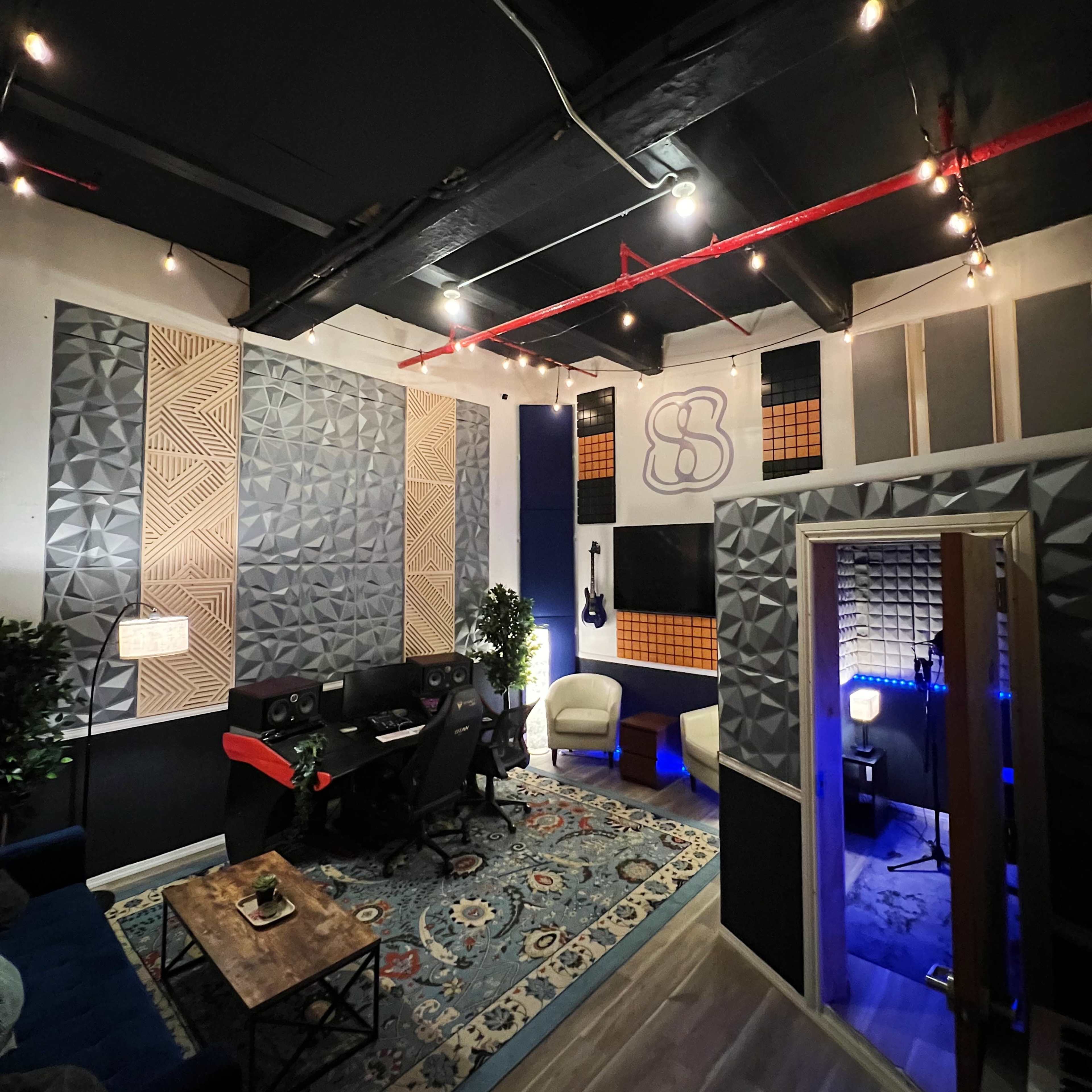 A music recording studio features soundproof walls, a control desk with computers, and cozy seating areas, illuminated by string lights.