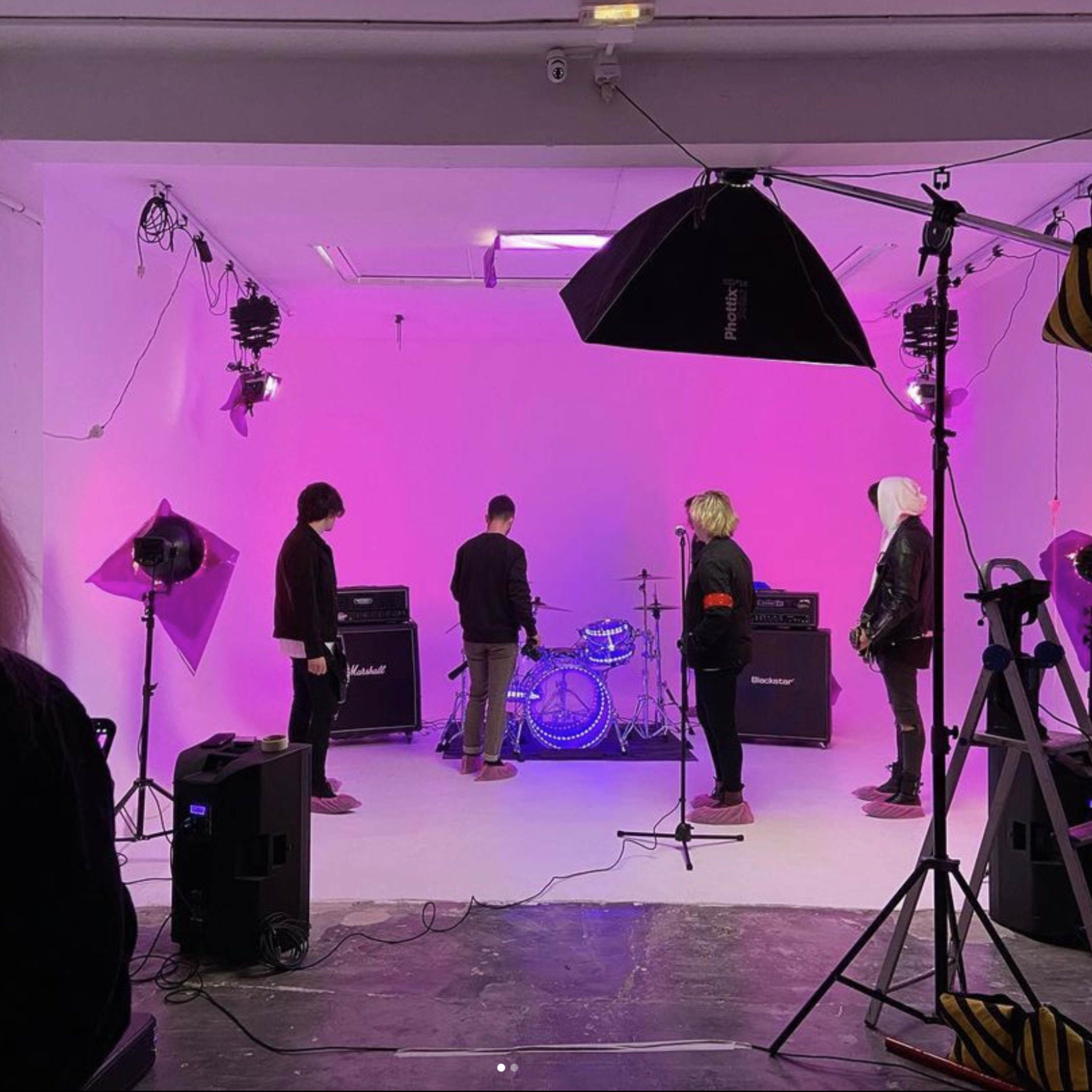 A band prepares to perform in a studio featuring pink lighting and various amplifiers arranged on a white stage.