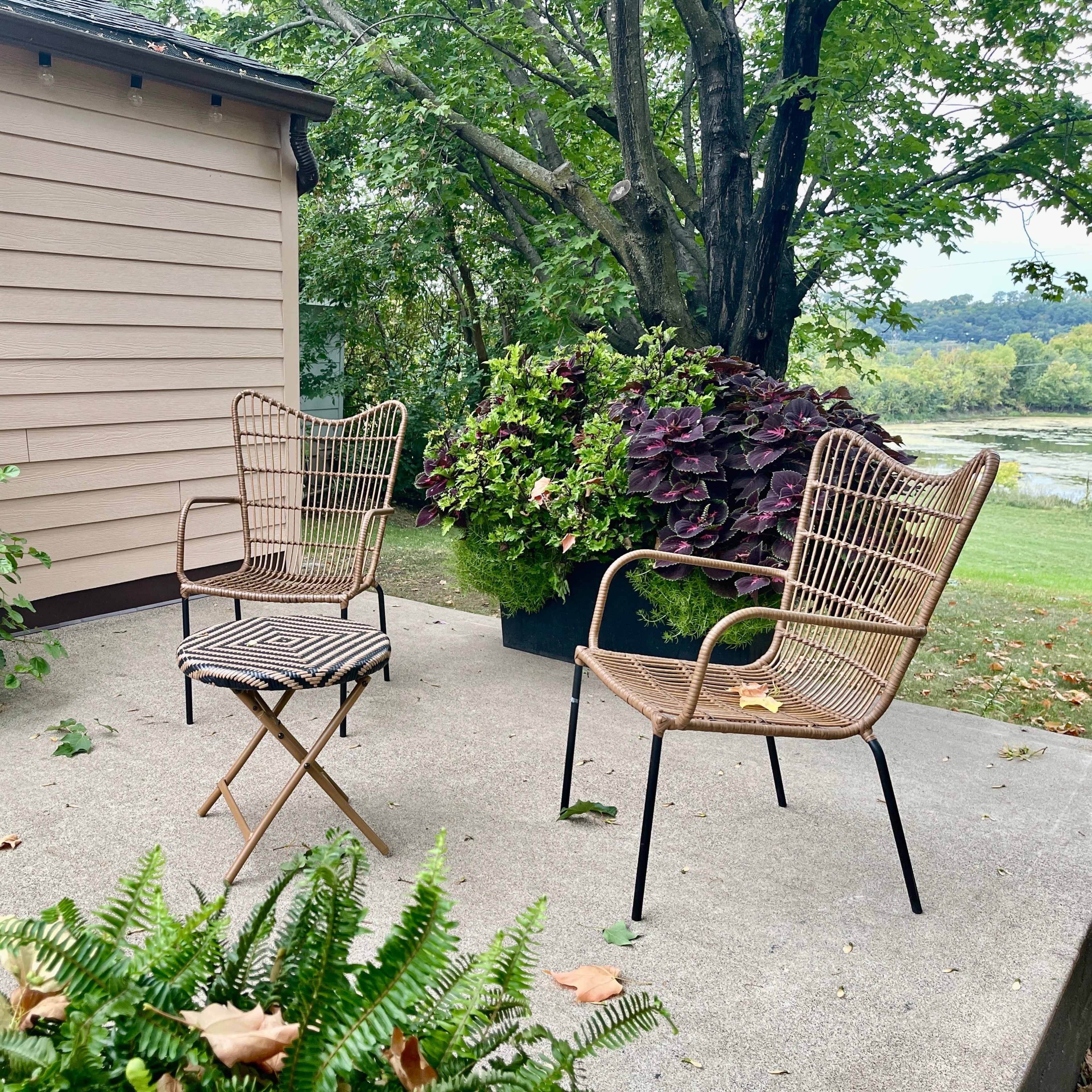 A patio area features two wicker chairs and a folding table surrounded by large potted plants.