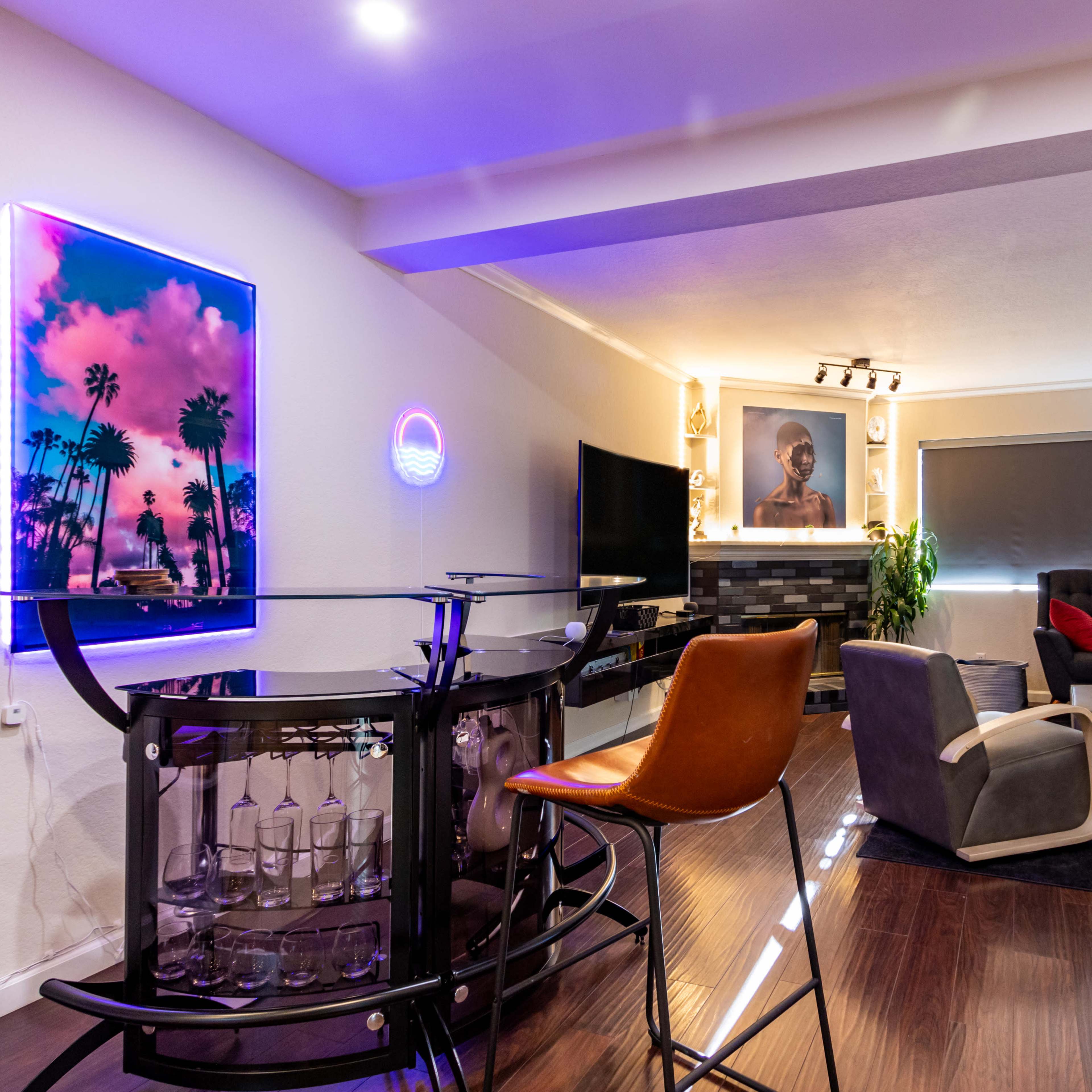 A modern living room featuring a seating area with a gray sofa, a black armchair, a bar with stools, and a decorative wall with artwork and a neon sign.