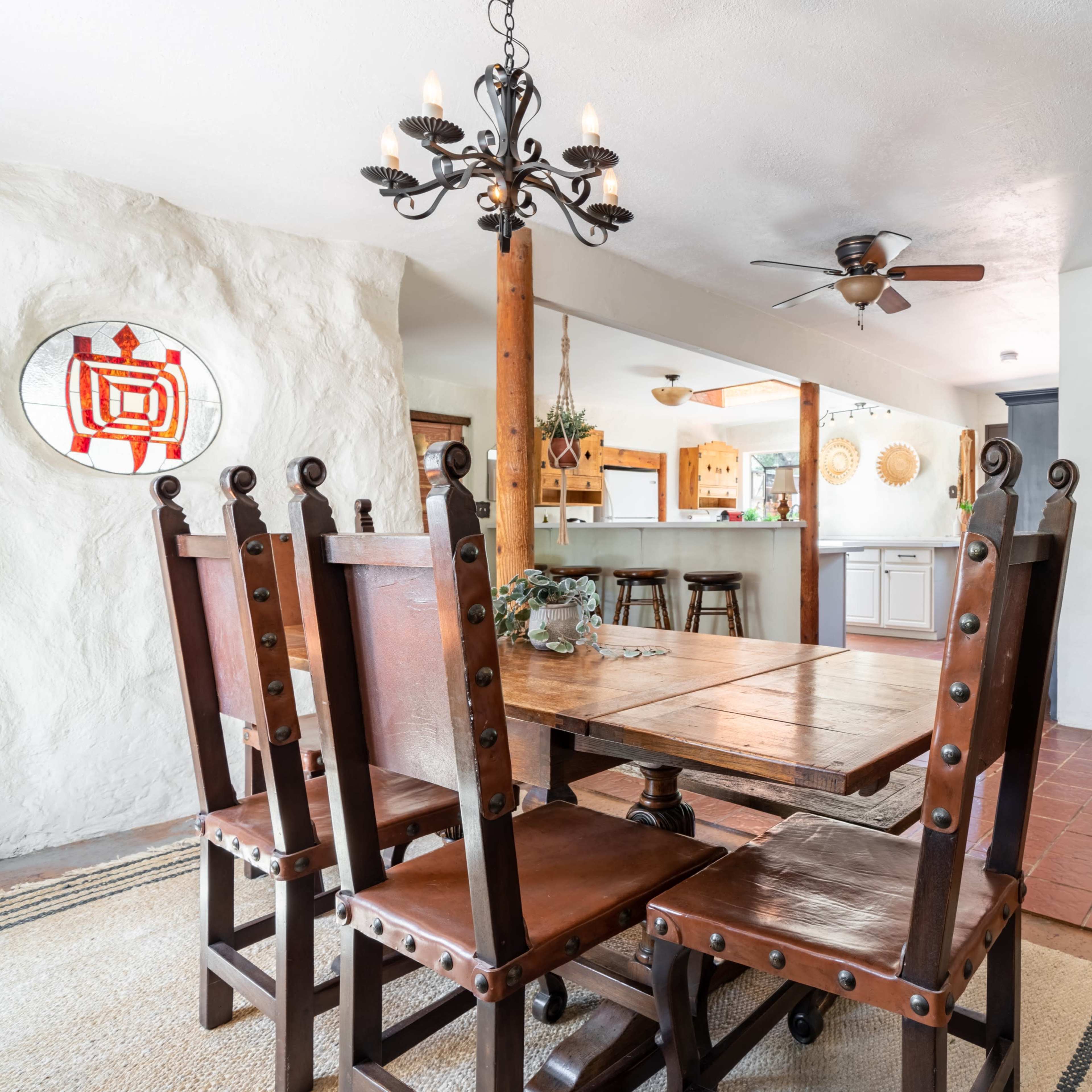 A dining area with a wooden table and chairs, a chandelier overhead, and a stove in the corner, with a view into a kitchen.