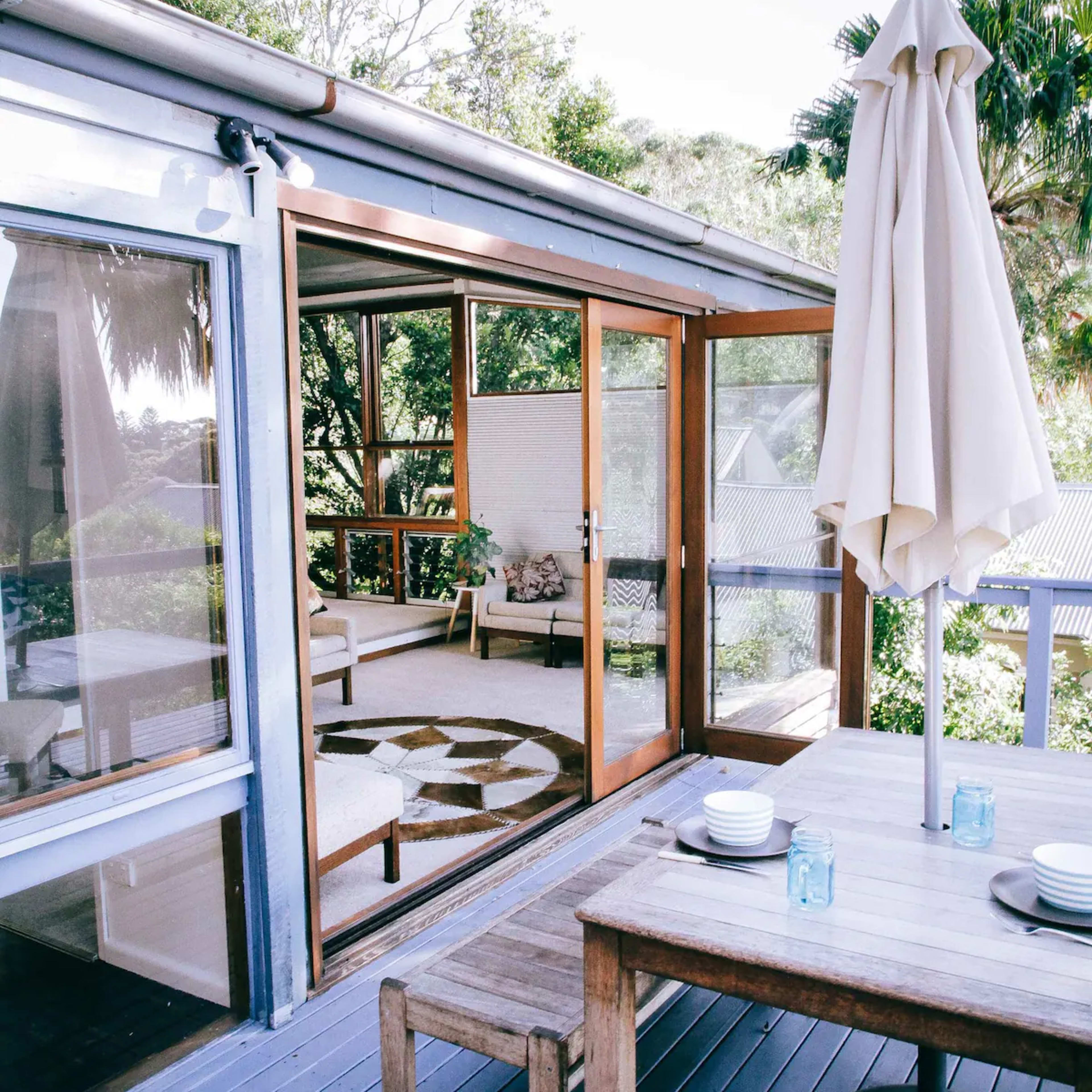 A patio area features a wooden table and chairs with a large umbrella overlooking a light-filled interior space through glass sliding doors.