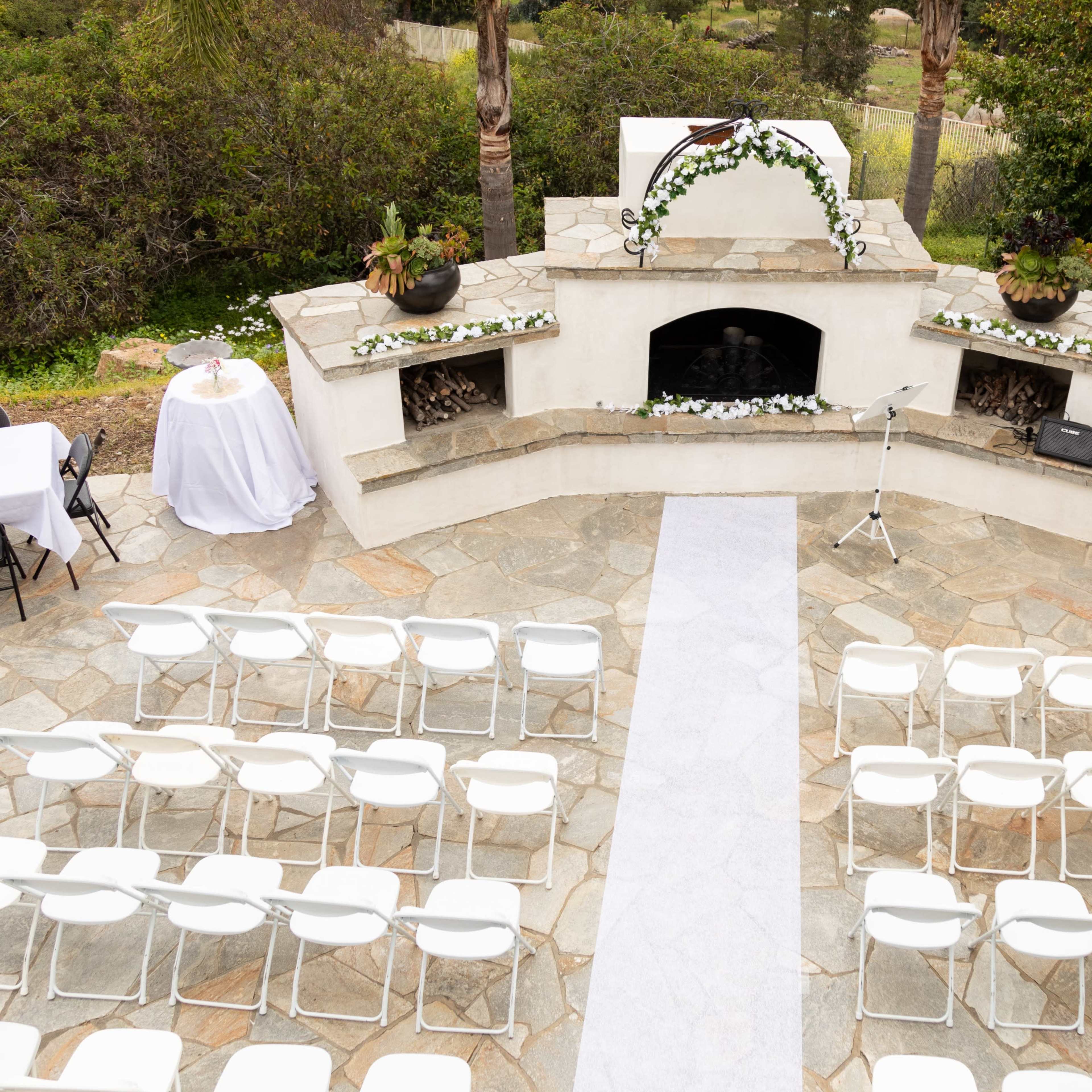 An outdoor wedding setup features white chairs arranged in rows facing a stone fireplace adorned with greenery and floral decorations.