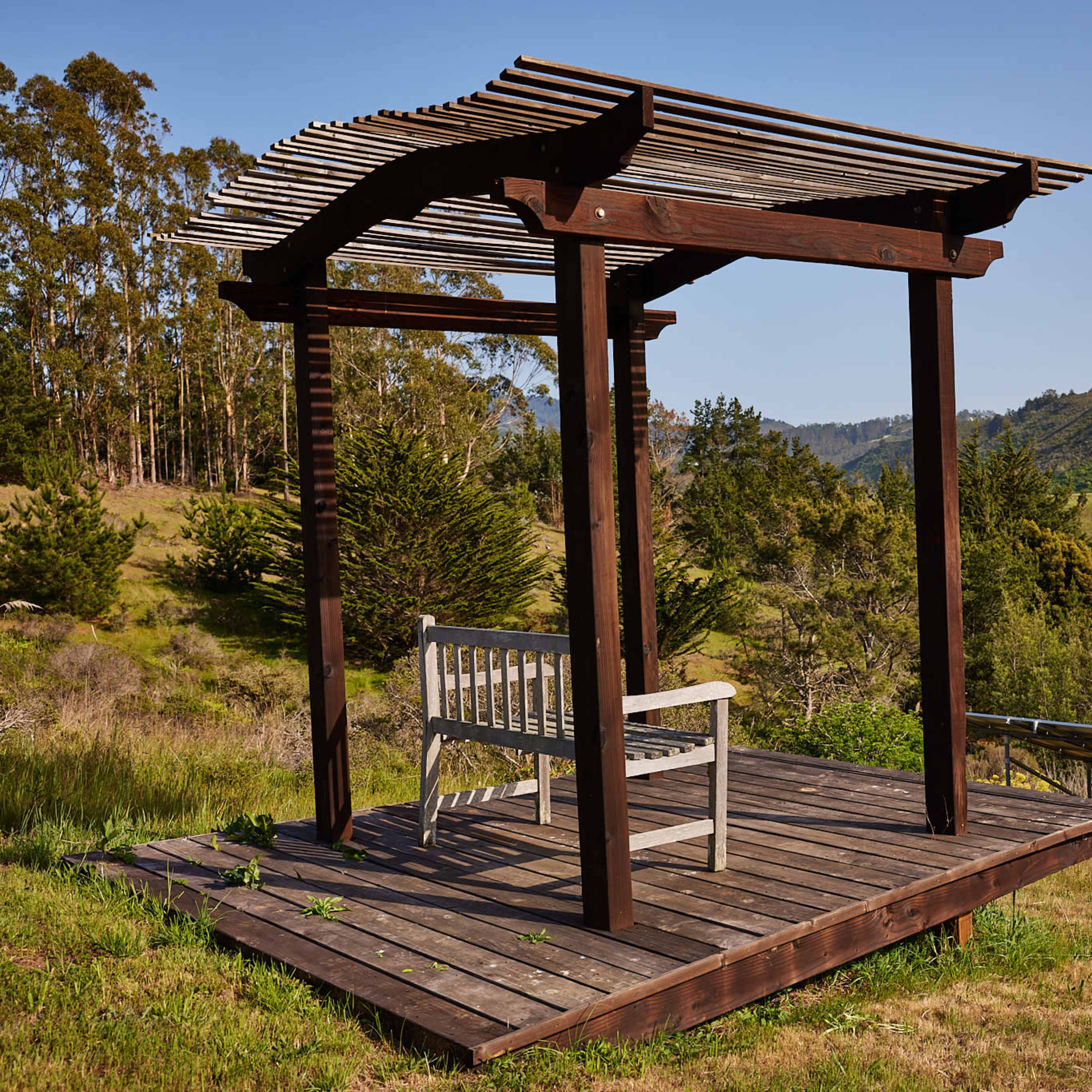 A wooden gazebo with a bench overlooks a grassy hillside and trees.