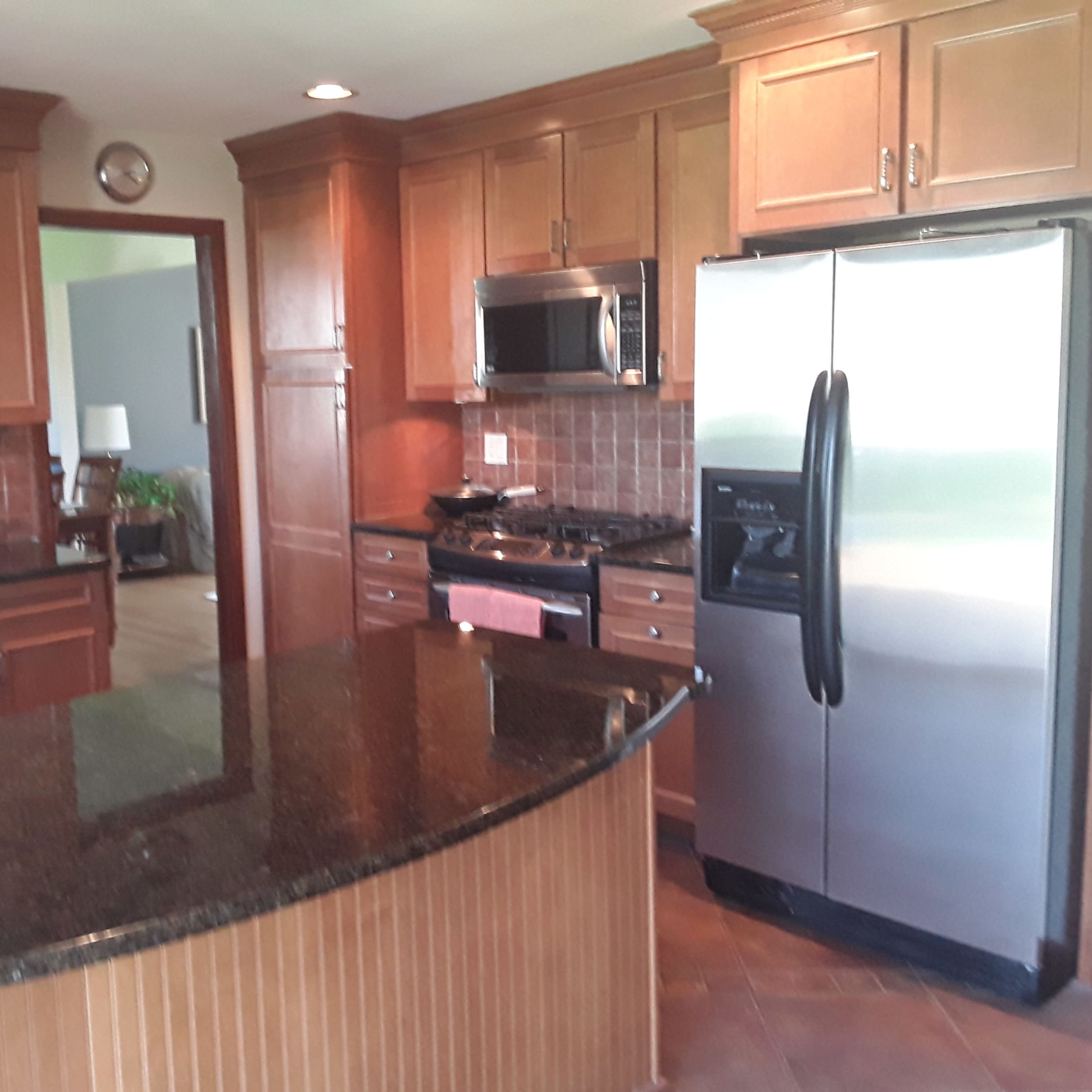 A modern kitchen with wooden cabinetry, a stainless steel refrigerator, and a black granite countertop, featuring a gas range and microwave built into the cabinetry.