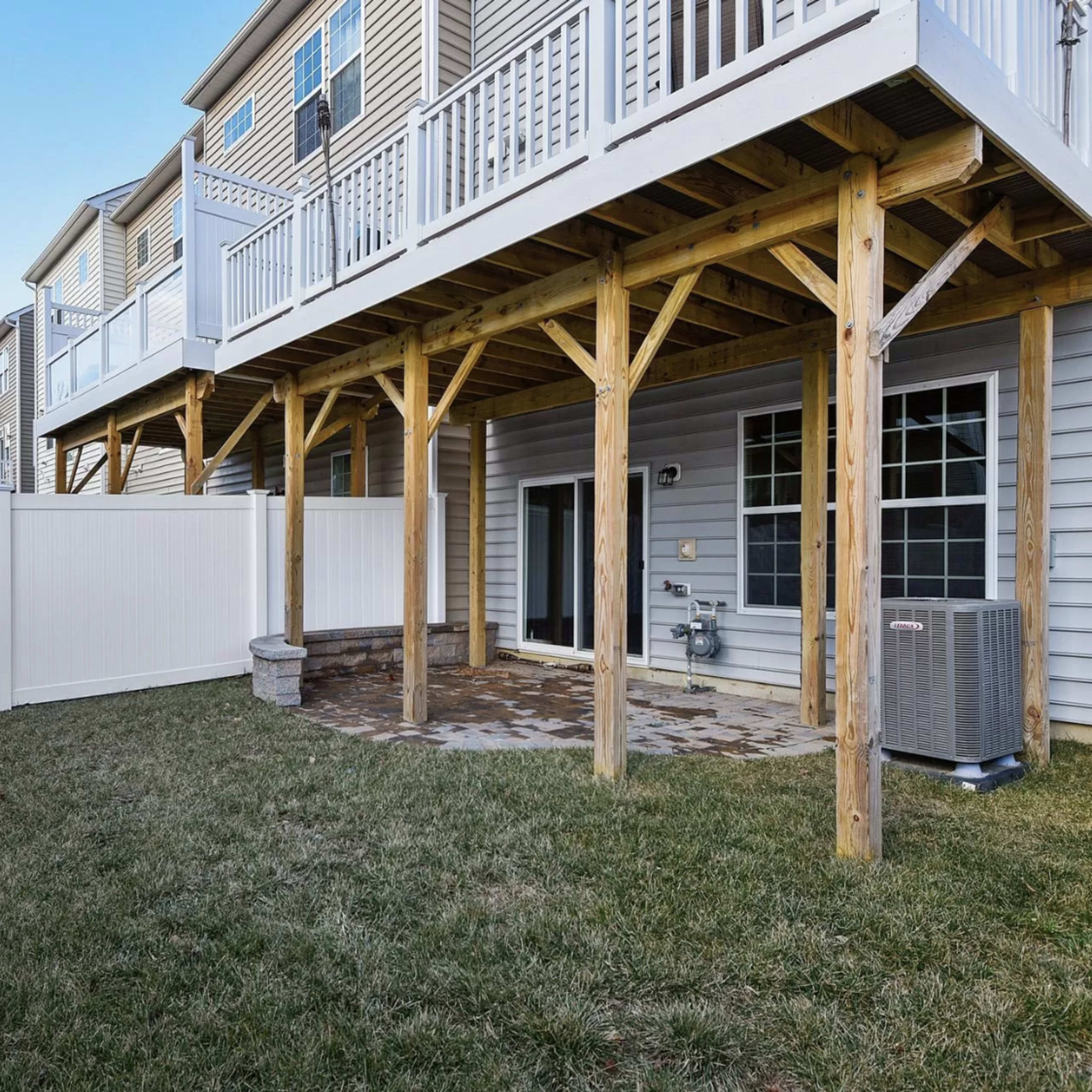 A backyard with a stone patio, surrounded by a white vinyl fence, beneath a deck supported by wooden posts.