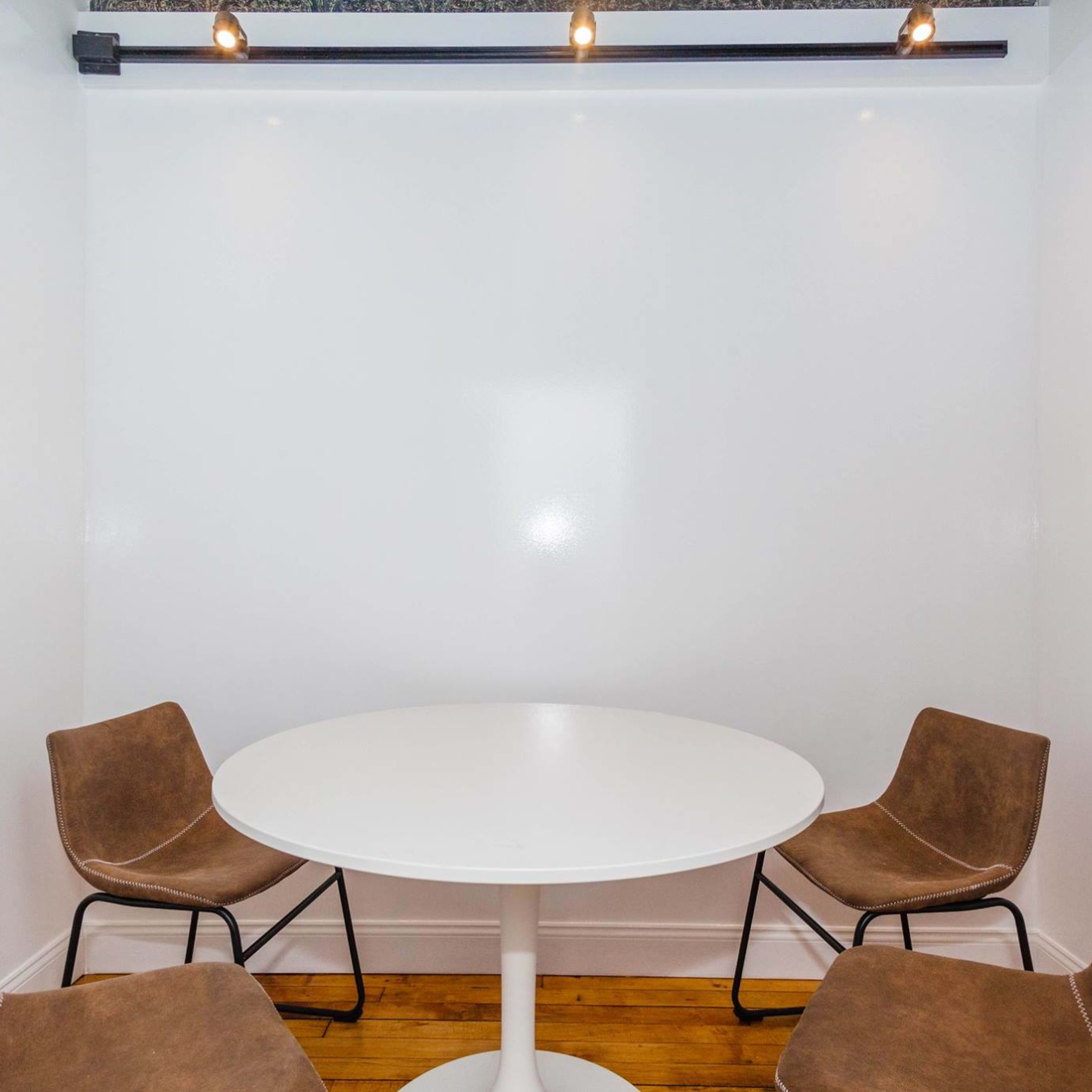 A small, empty room features a white circular table surrounded by four brown chairs.