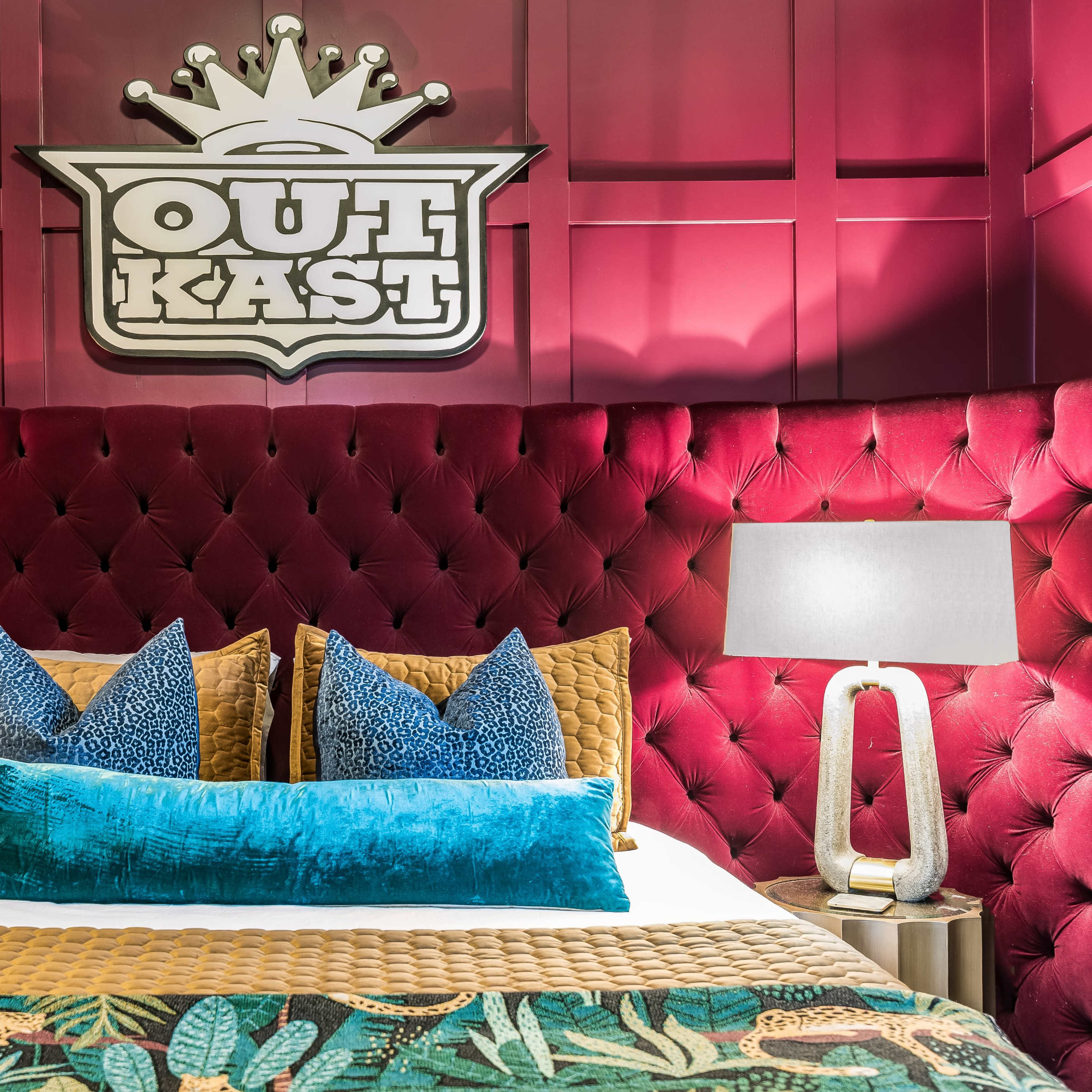 A stylized hotel room with a plush red upholstered headboard featuring the "OutKast" logo, flanked by modern table lamps and decorative pillows on the bed.