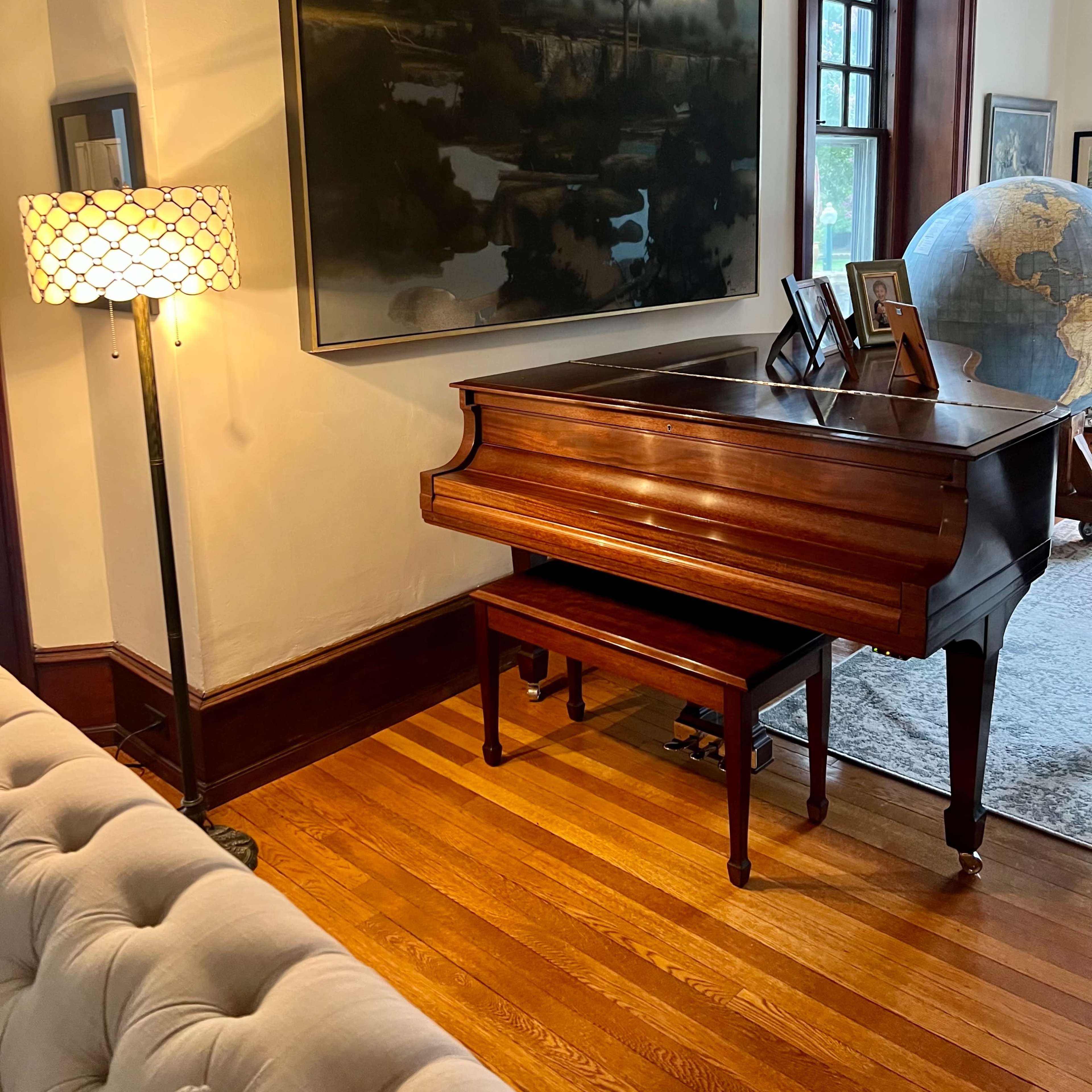 A wooden piano is positioned next to a floor lamp and a large painting in a well-lit room.