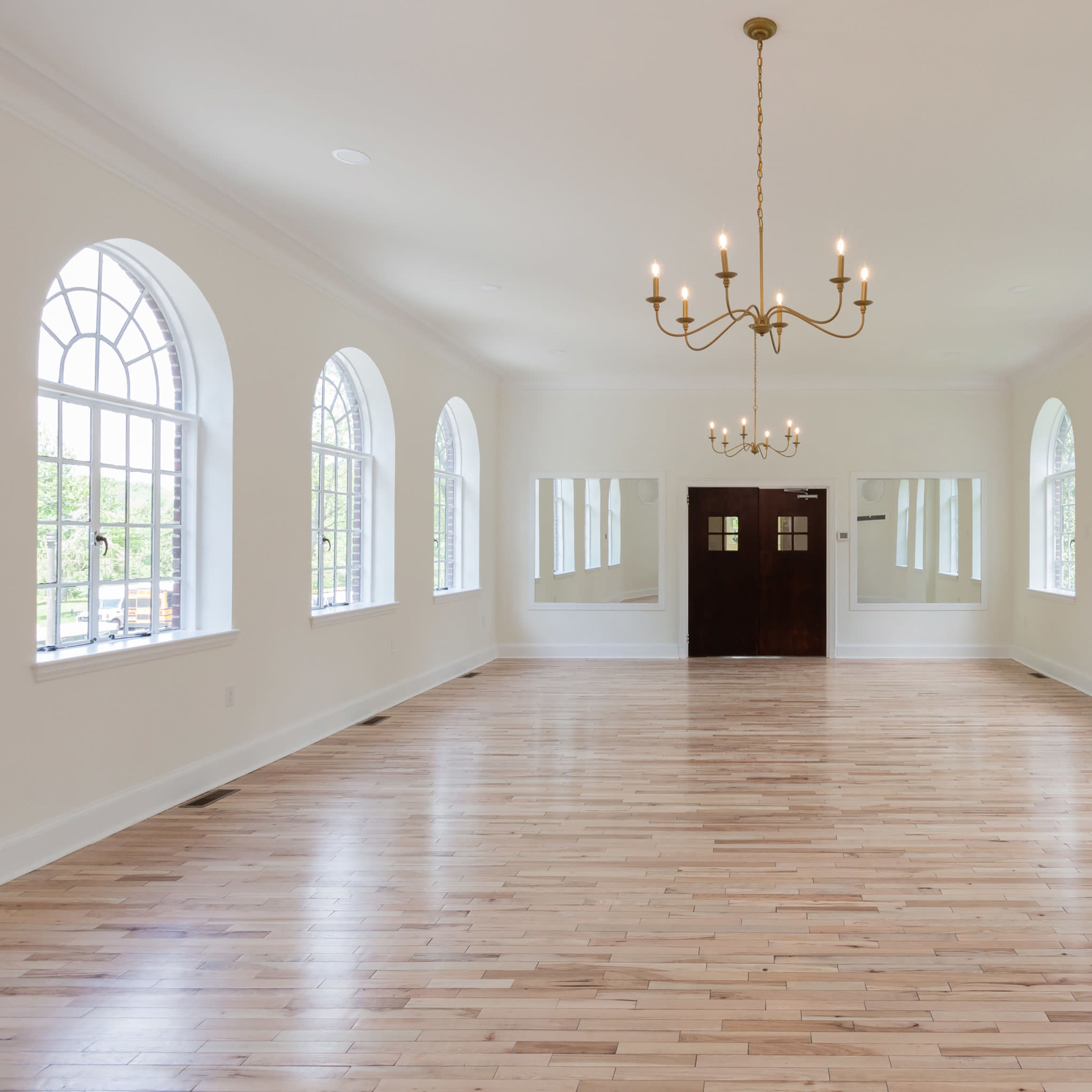 A spacious, well-lit room with hardwood floors, large arched windows, and a chandelier overhead, leading to a set of double doors.