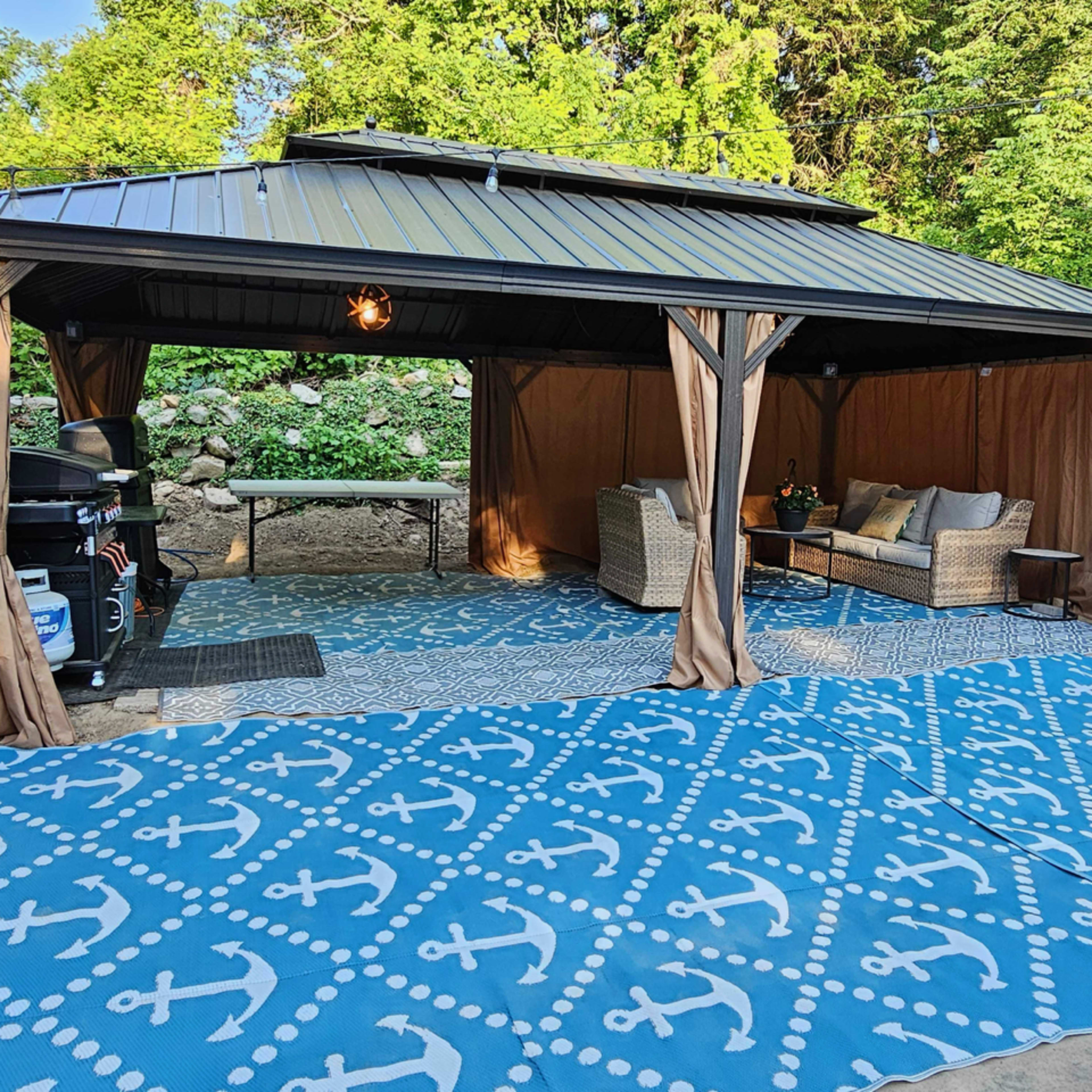 A covered outdoor seating area features beige furniture and a blue anchor-patterned rug, surrounded by greenery.