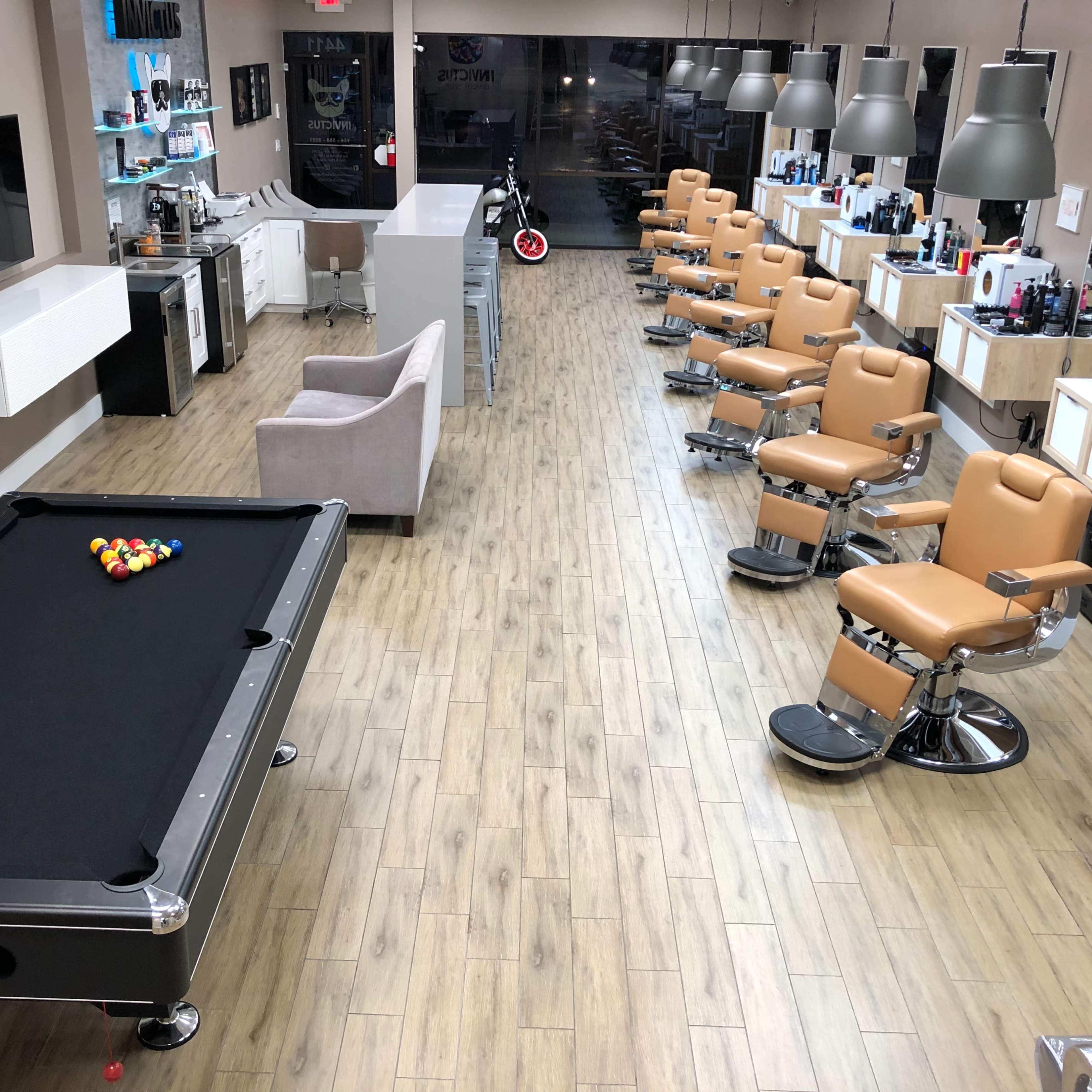 A spacious barbershop with several styling chairs lined up, a pool table in the foreground, and various grooming products displayed on counters.