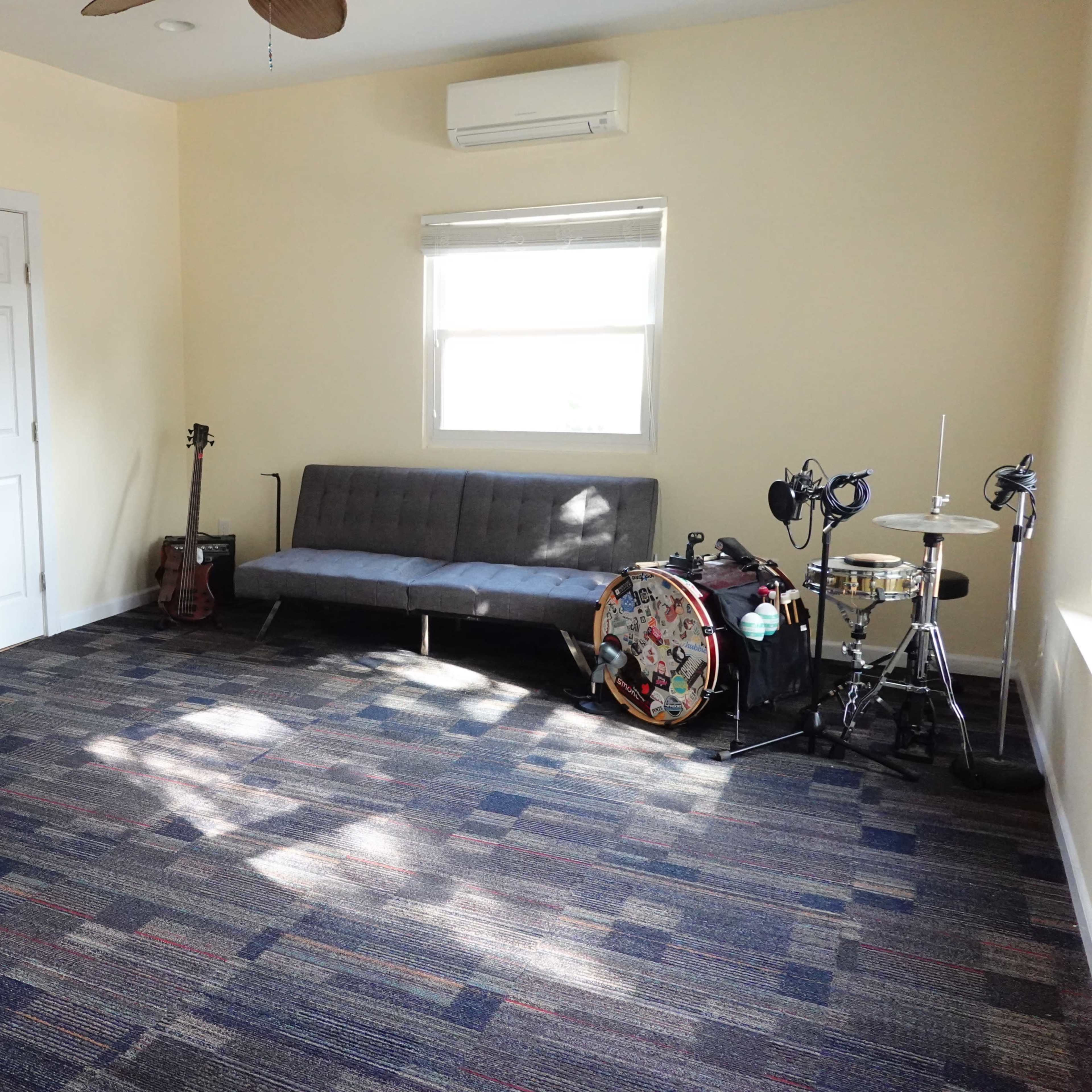 The room features a futon, a keyboard, a guitar, and a drum set, with a window letting in natural light.