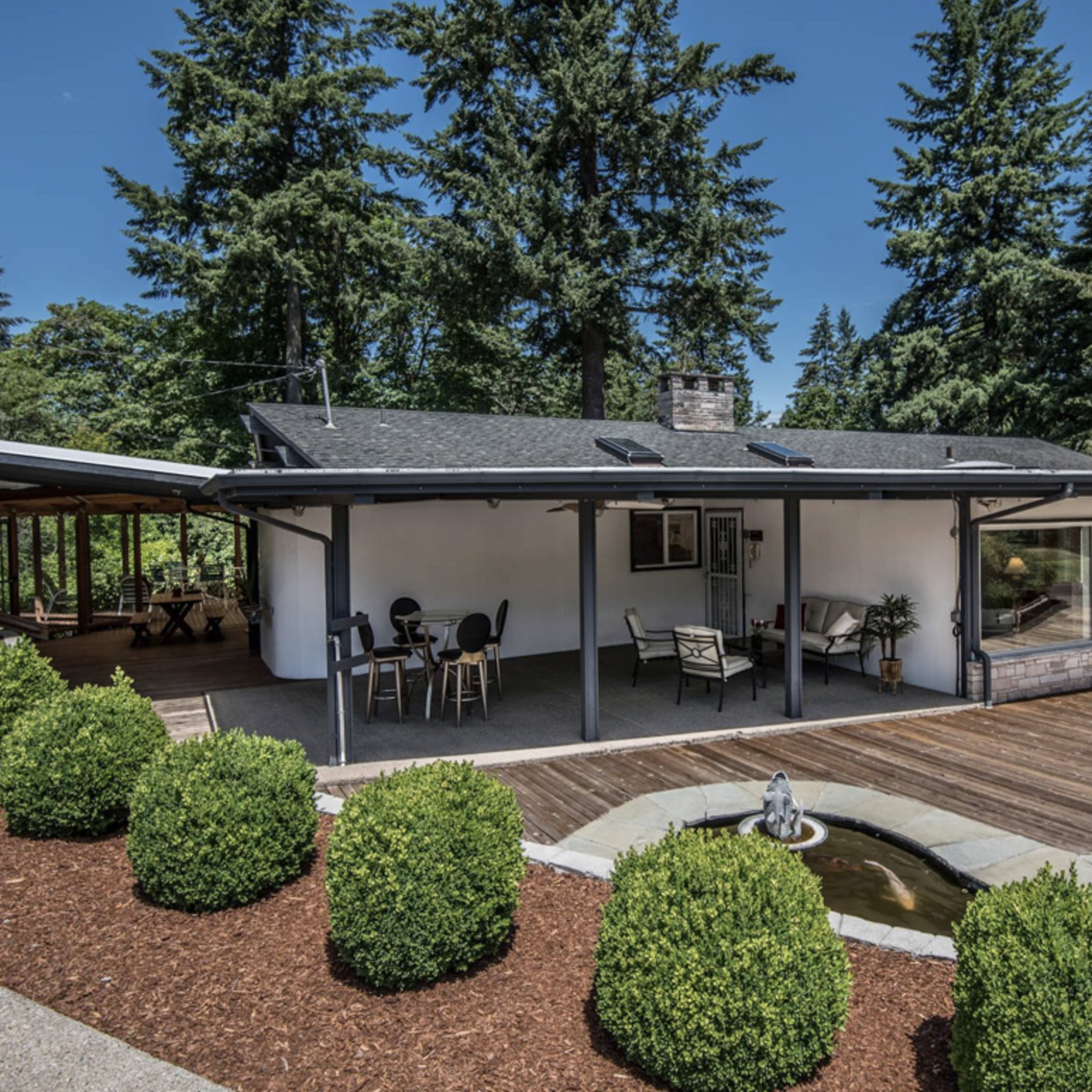A modern house with a wooden deck and outdoor seating area is surrounded by neatly trimmed shrubs and tall trees.