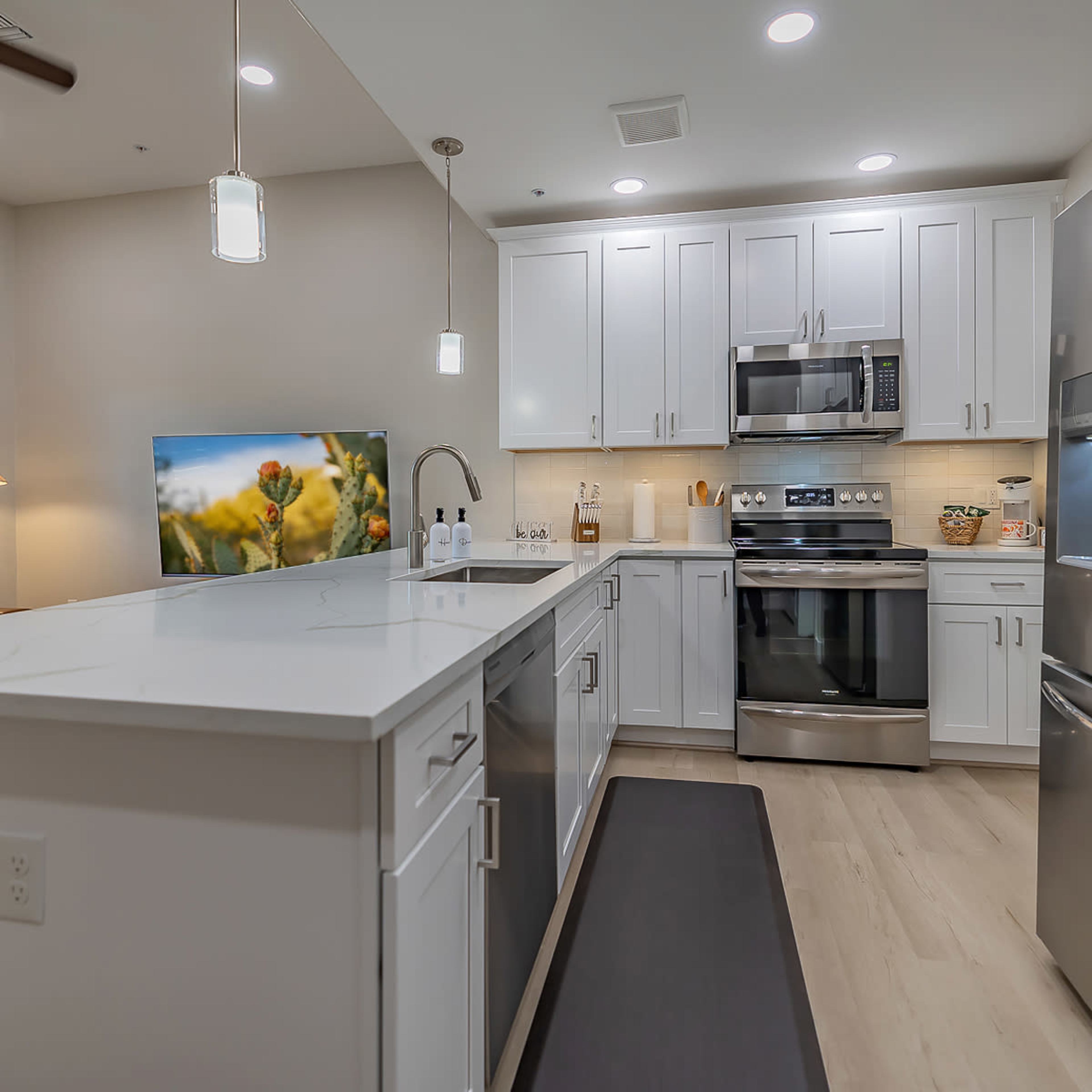 A modern kitchen features white cabinetry, stainless steel appliances, and an island with a sink, opening to a living area with a leather couch and a television.