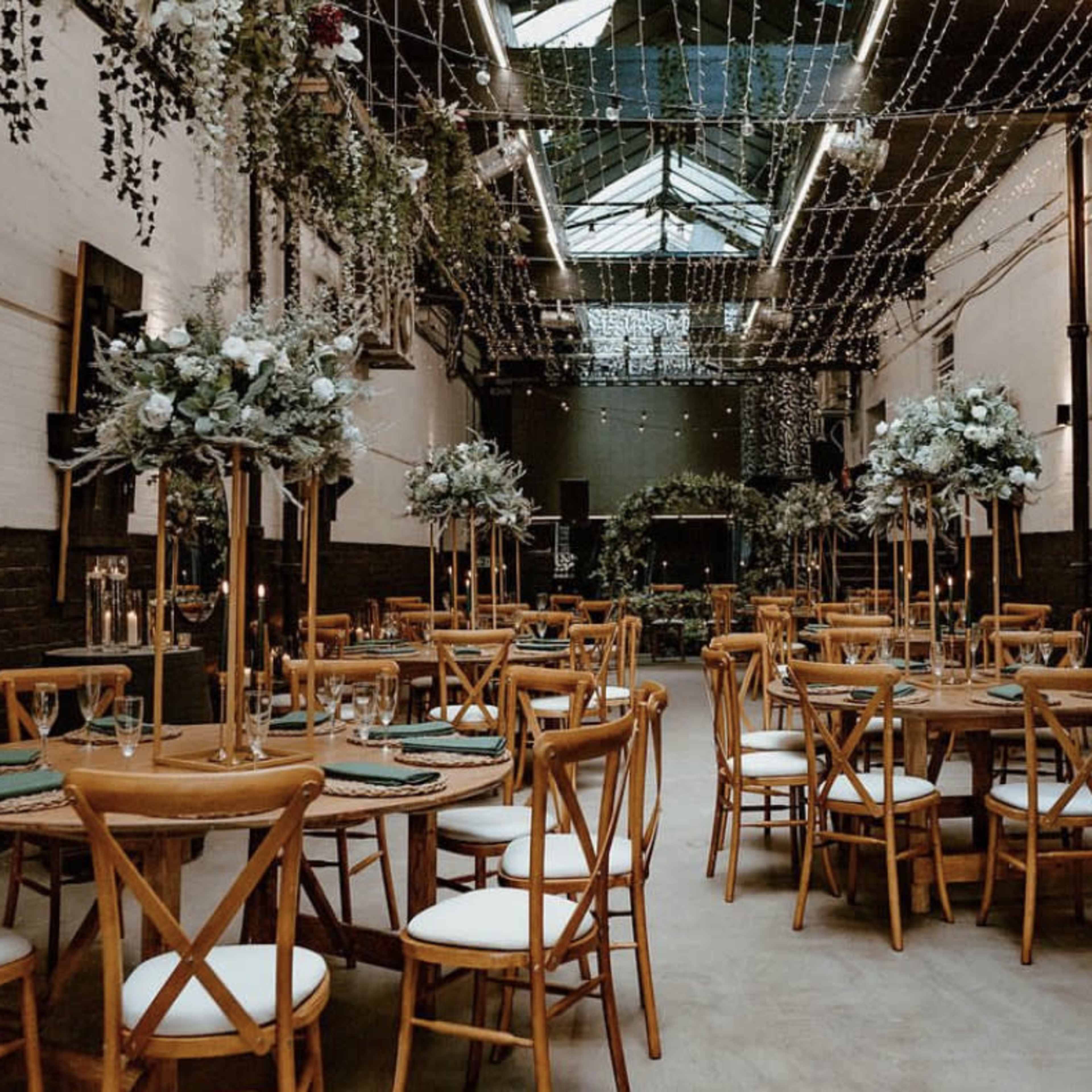 A spacious event hall features wooden tables set with greenery and candles, illuminated by string lights hanging from the ceiling.