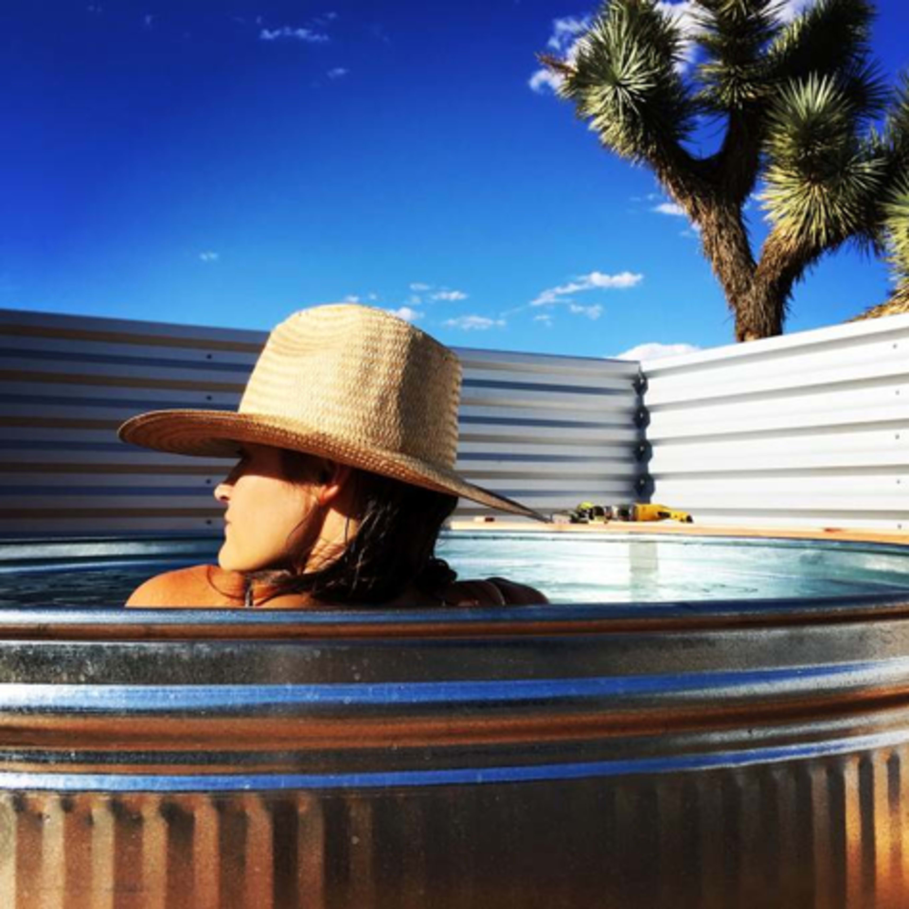 A person wearing a straw hat relaxes in a metal hot tub against a backdrop of clear blue sky and a Joshua tree.