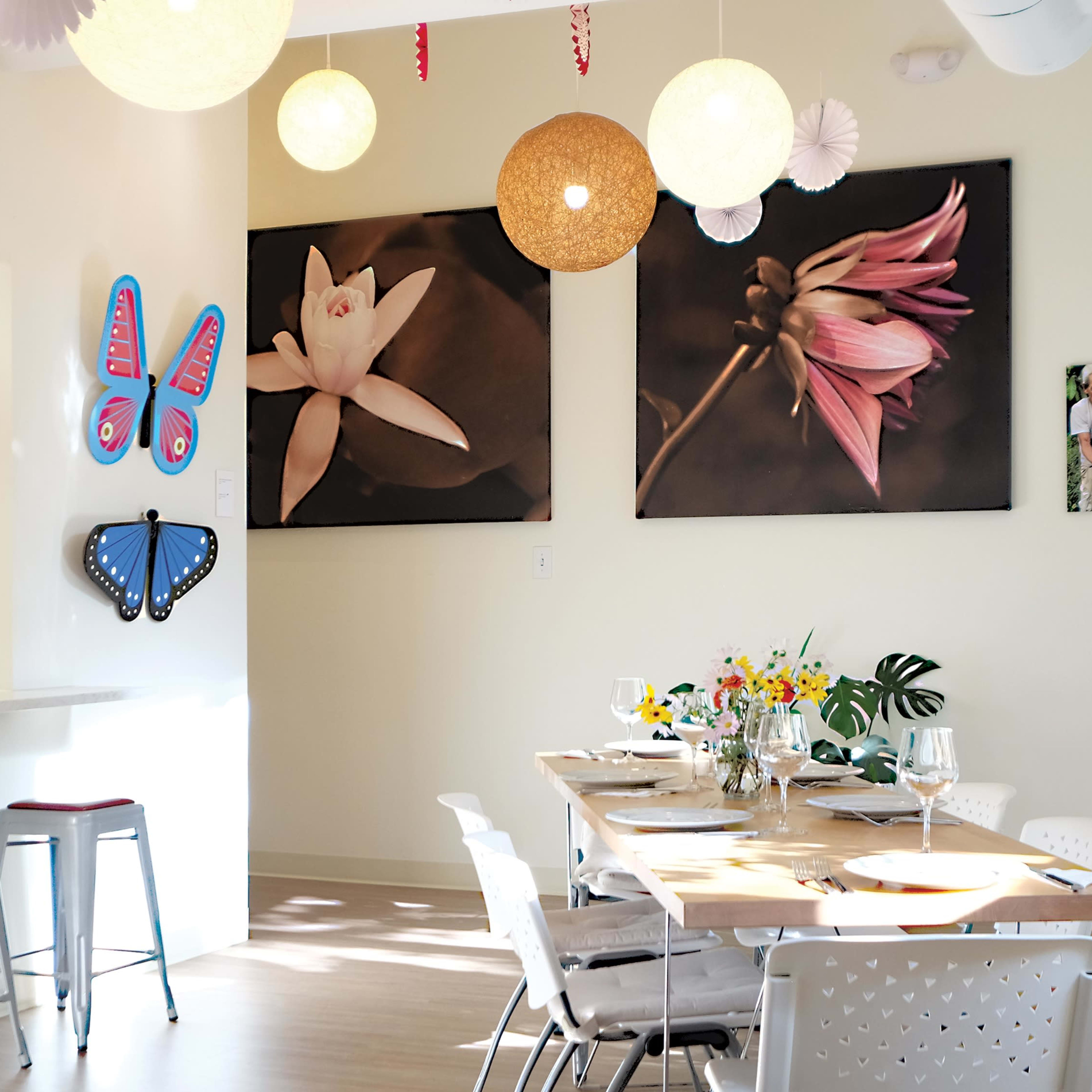 A bright dining area features a wooden table set for a meal, surrounded by white chairs, with colorful wall decorations and large floral photographs.