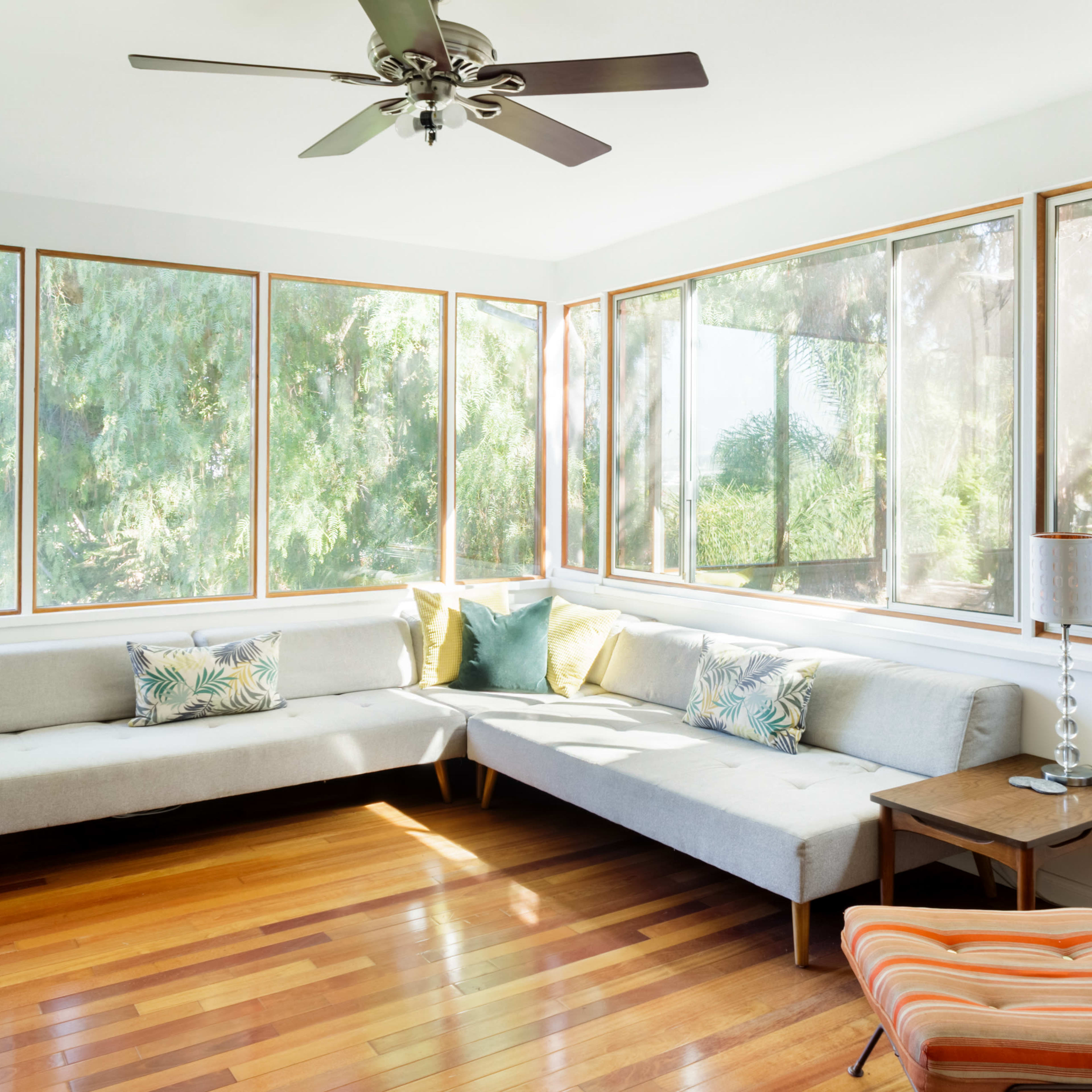 A bright living room features large windows, a sectional sofa, and wooden flooring, accented by a ceiling fan and side tables.