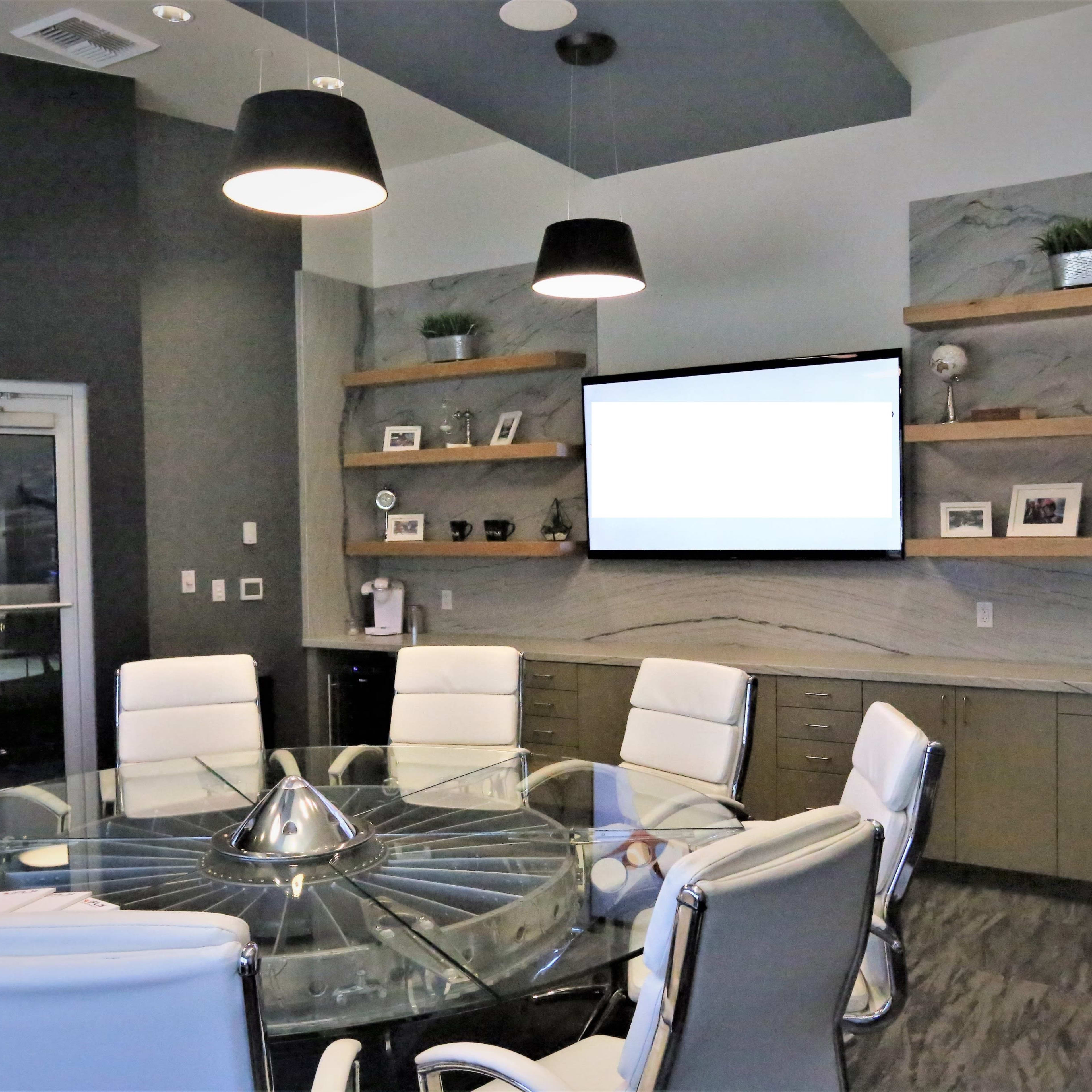A modern conference room featuring a circular glass table surrounded by white leather chairs, with wall-mounted shelves displaying decorative items and a large screen at the front.