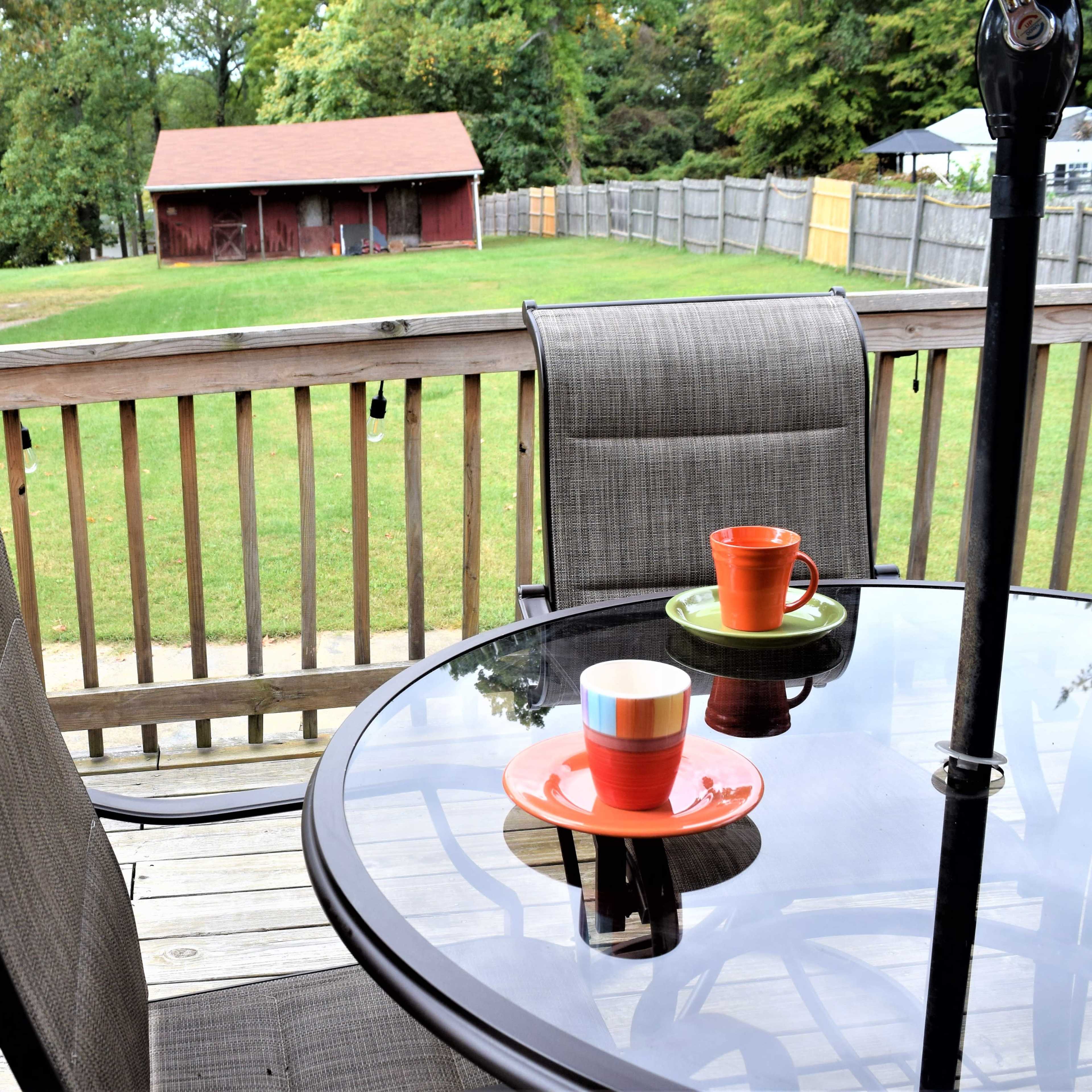 A glass-topped table with two colorful mugs and plates sits on a wooden deck overlooking a grassy yard and a red shed in the background.