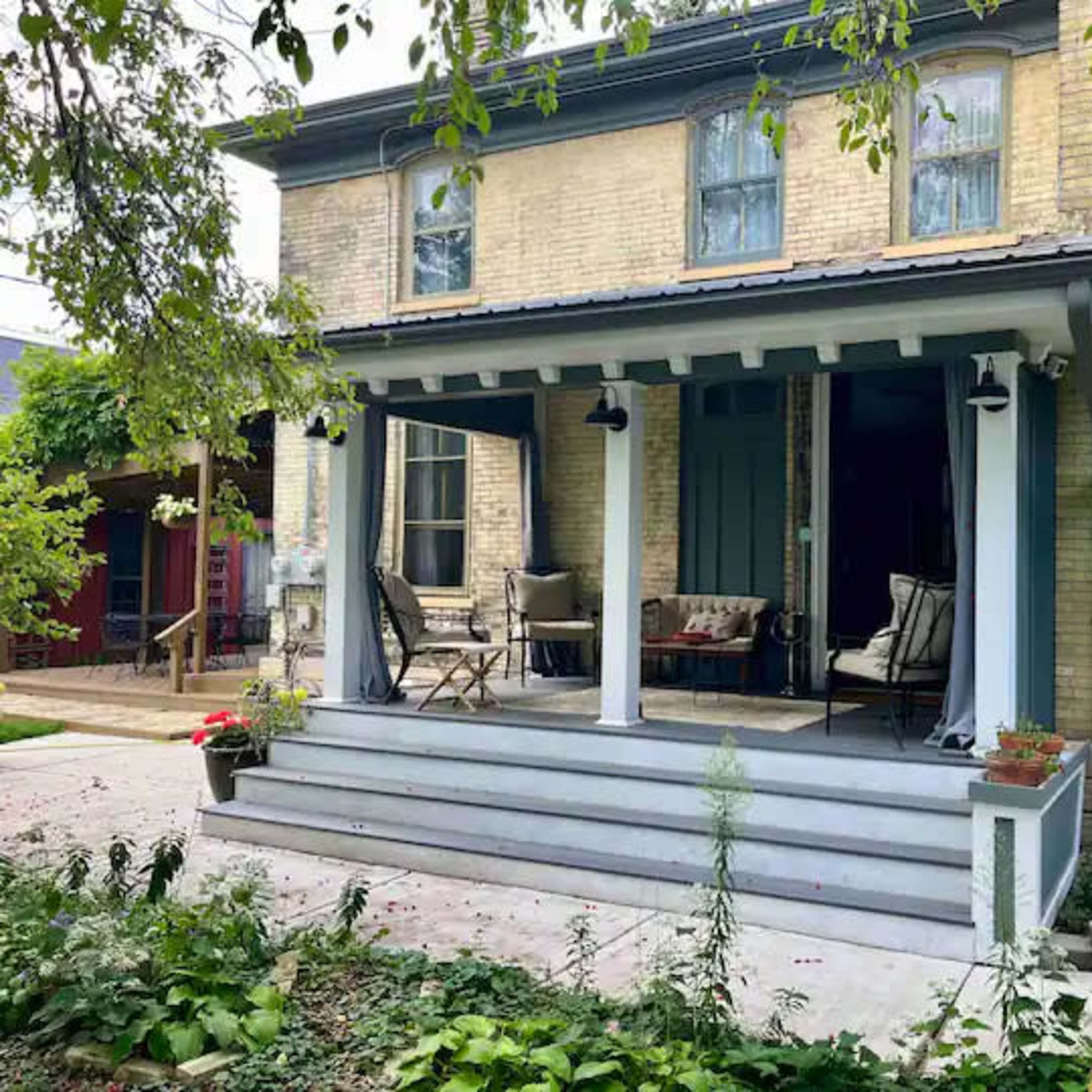 A two-story house with a porch featuring seating and potted plants, surrounded by greenery and a paved pathway.