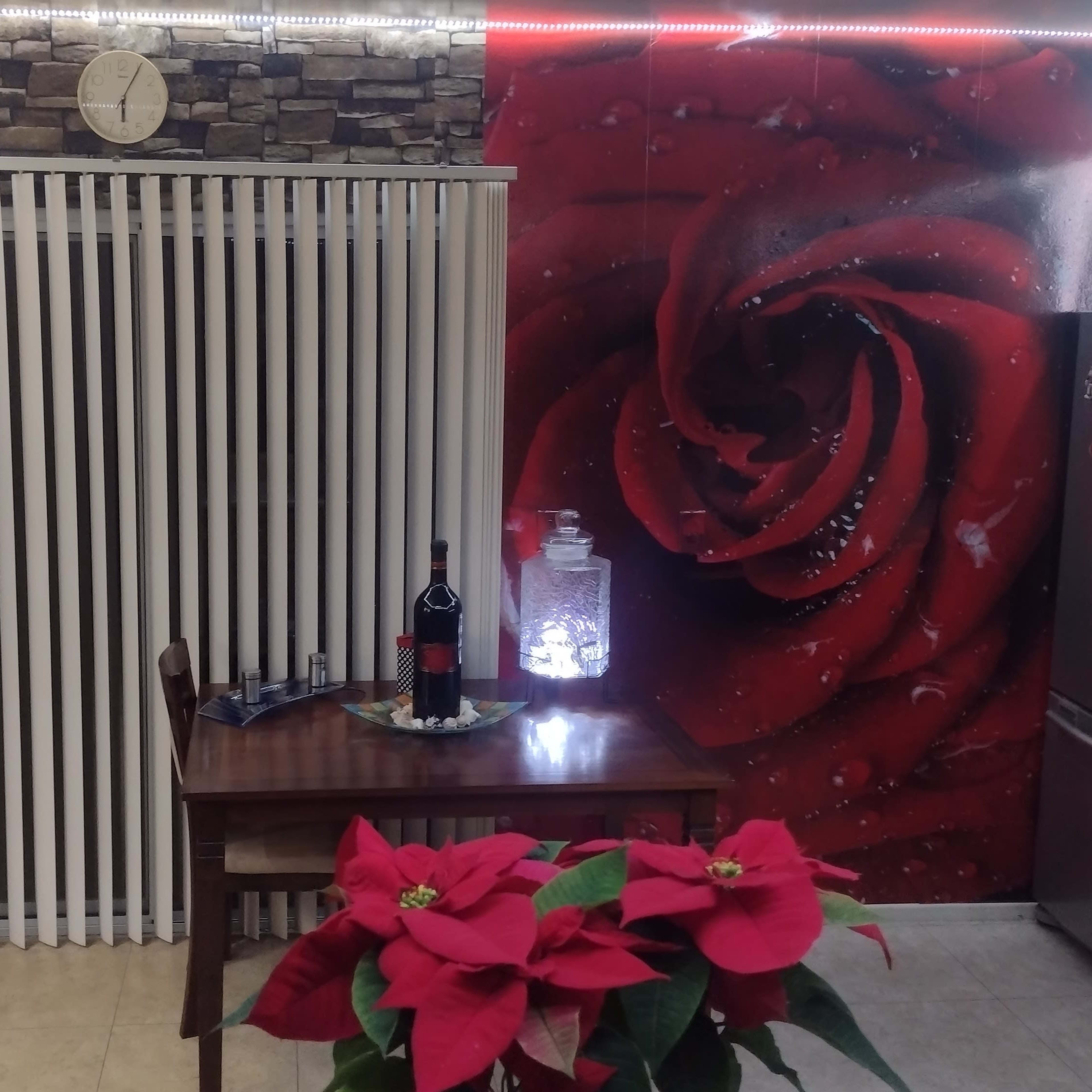 A dining area with a table set for drinks, a decorative wall featuring a large red rose, and a window with vertical blinds.