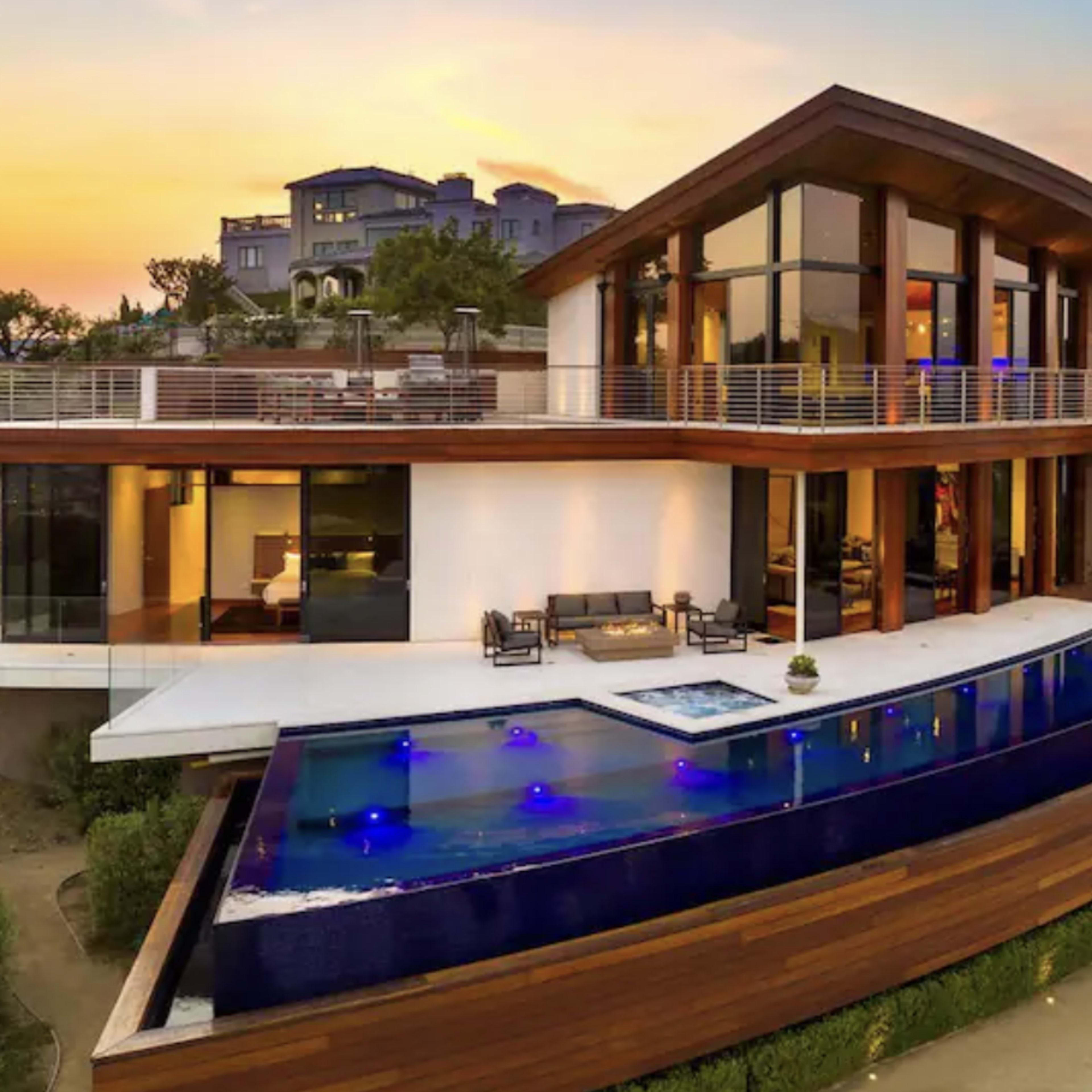 A modern house with large windows, featuring a sleek swimming pool that extends toward the edge of a hillside, all set against a sunset sky.