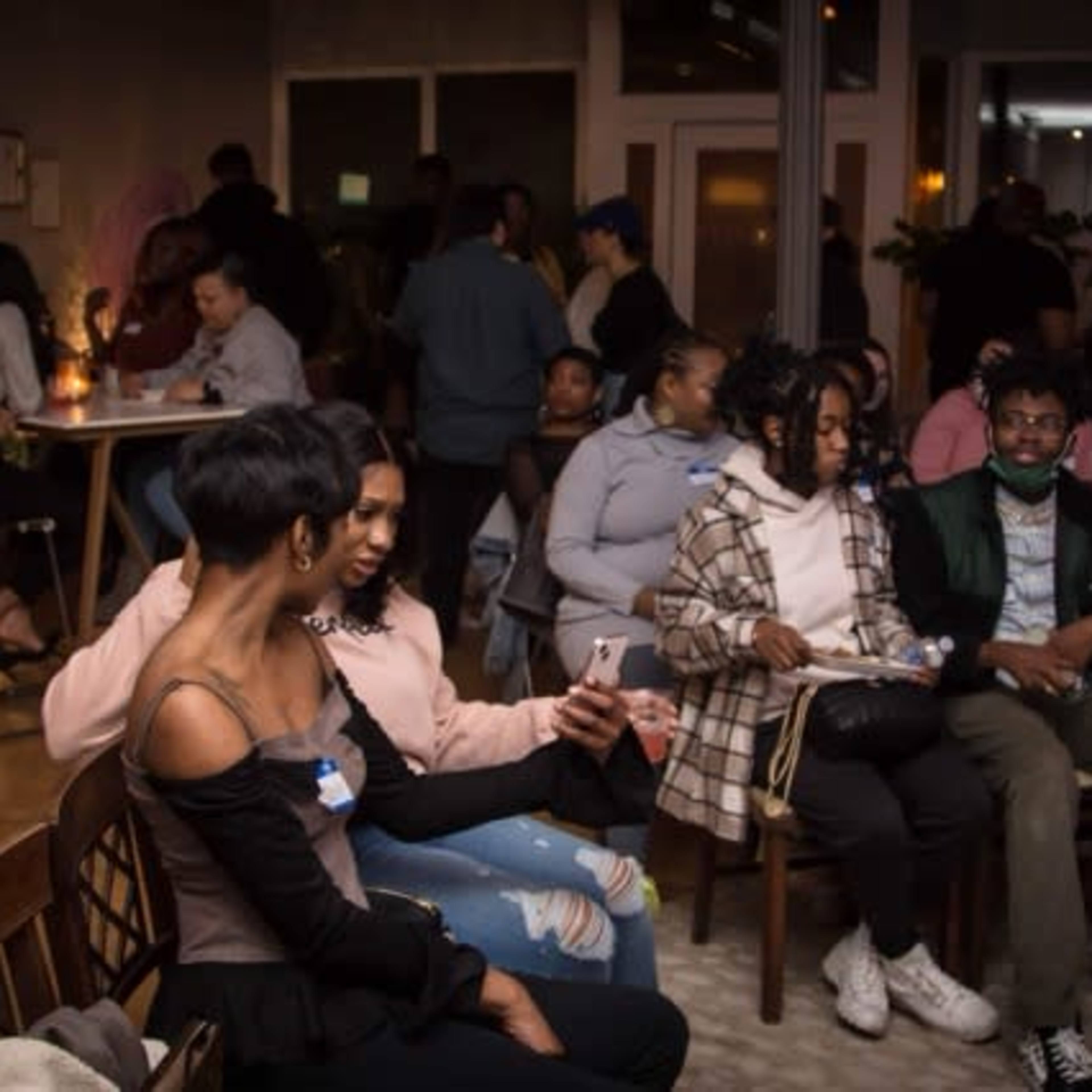 A group of people socializes in a lively indoor venue with tables and chairs, while some individuals are engaged in conversation and others are using their phones.