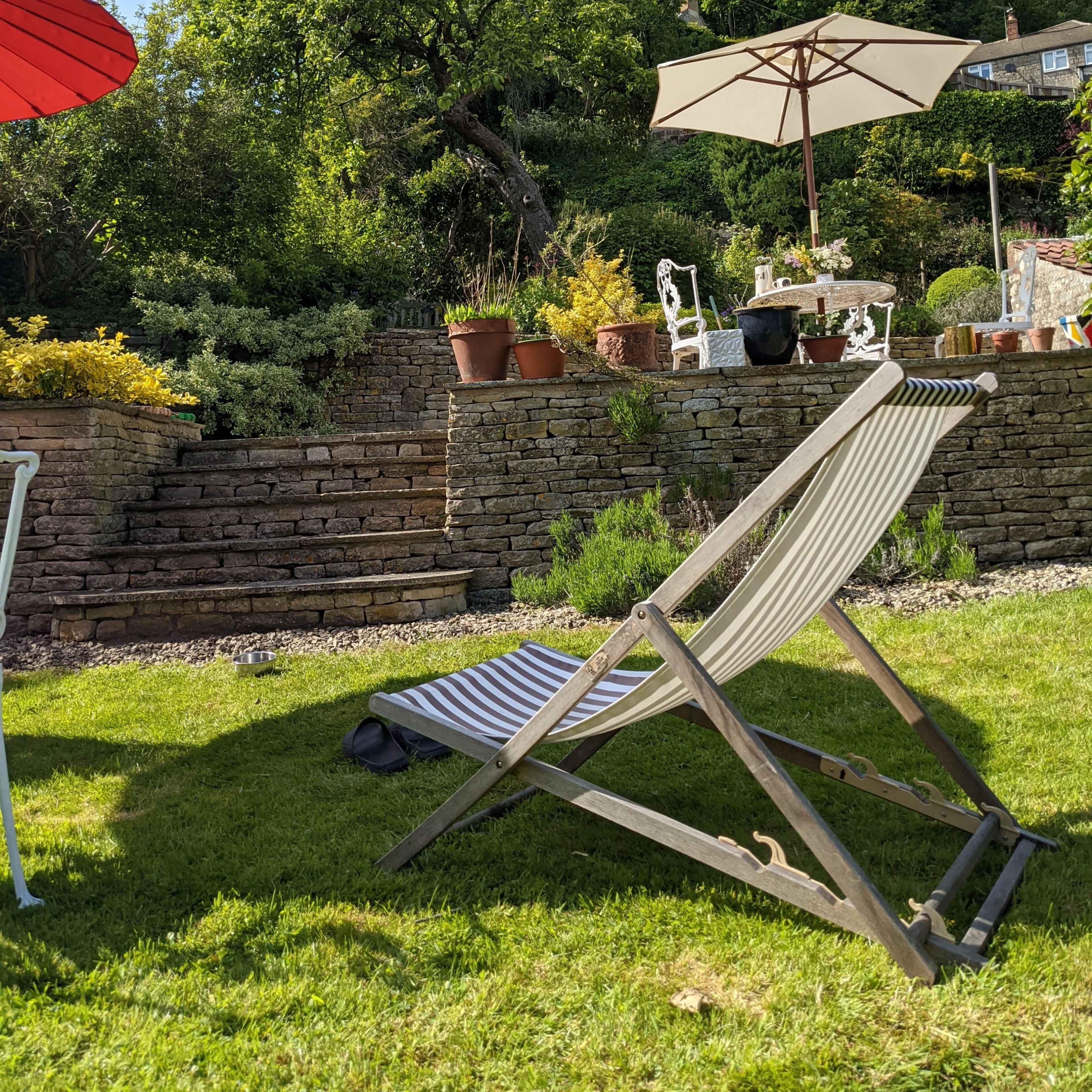 A wooden lounge chair with striped fabric rests on green grass in a garden with a stone stairway and potted plants.