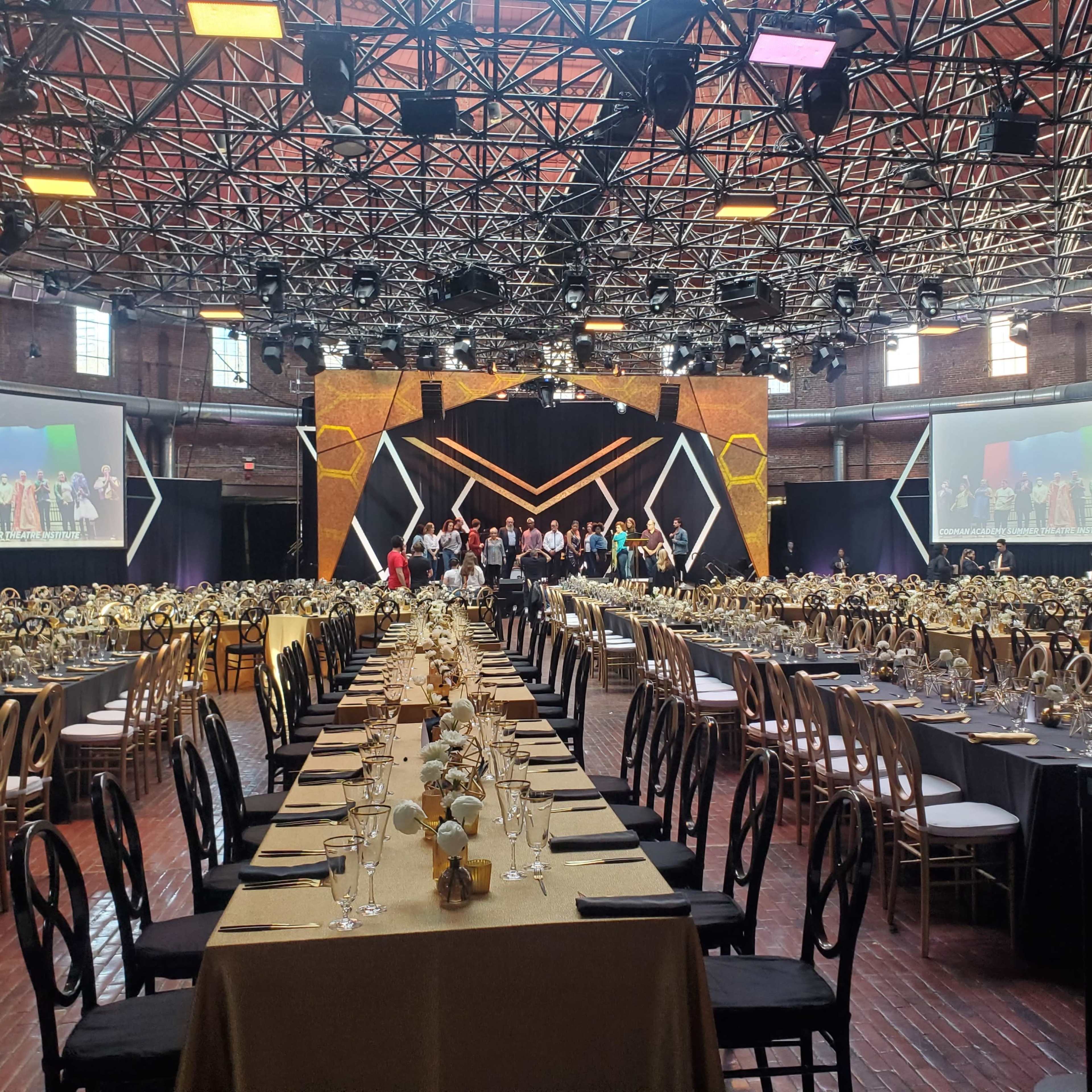 A large banquet hall is set up with elegantly arranged tables, and a stage in the background displays a group of people, with projection screens on either side.