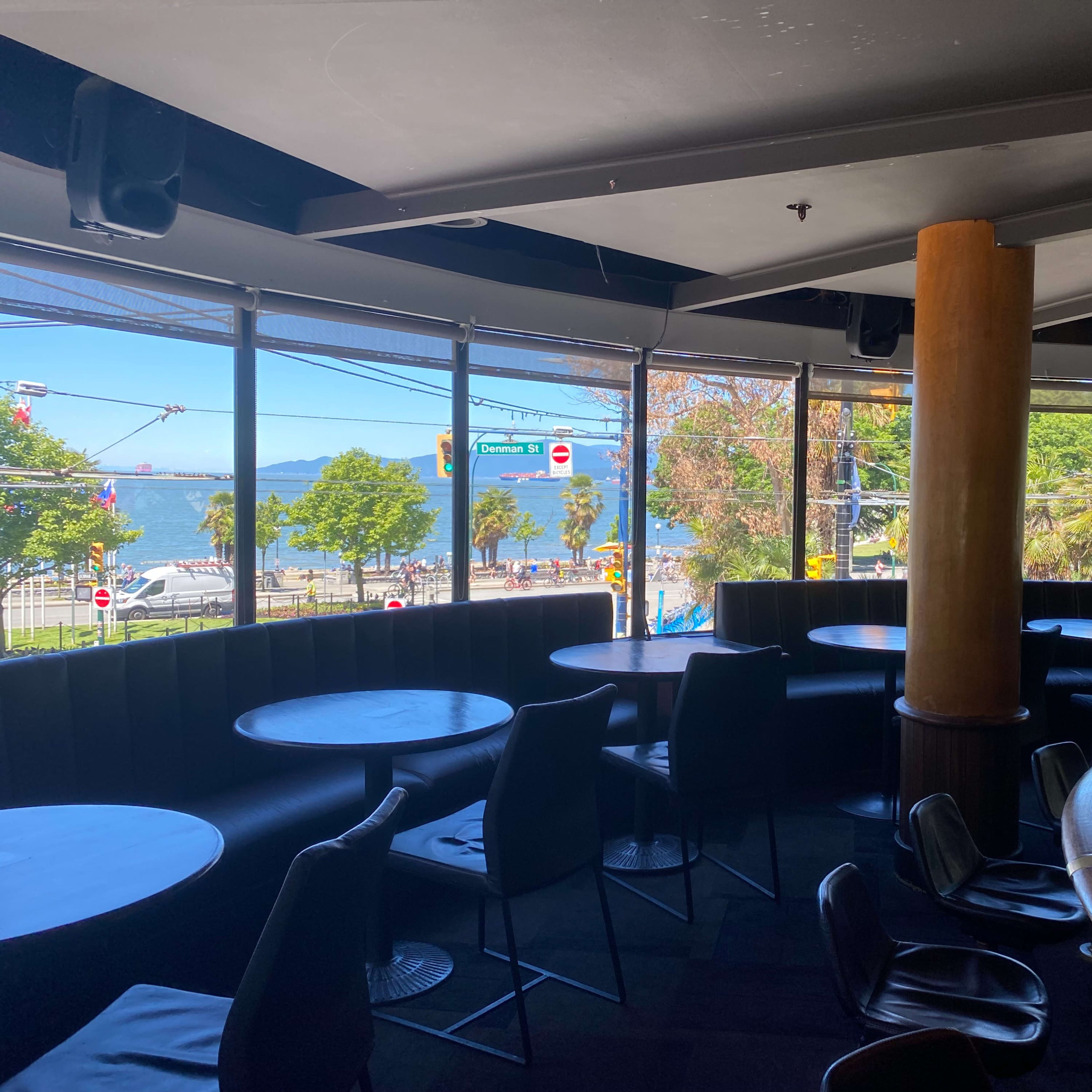 The interior of a bar features empty tables and chairs with large windows overlooking a sunny waterfront.