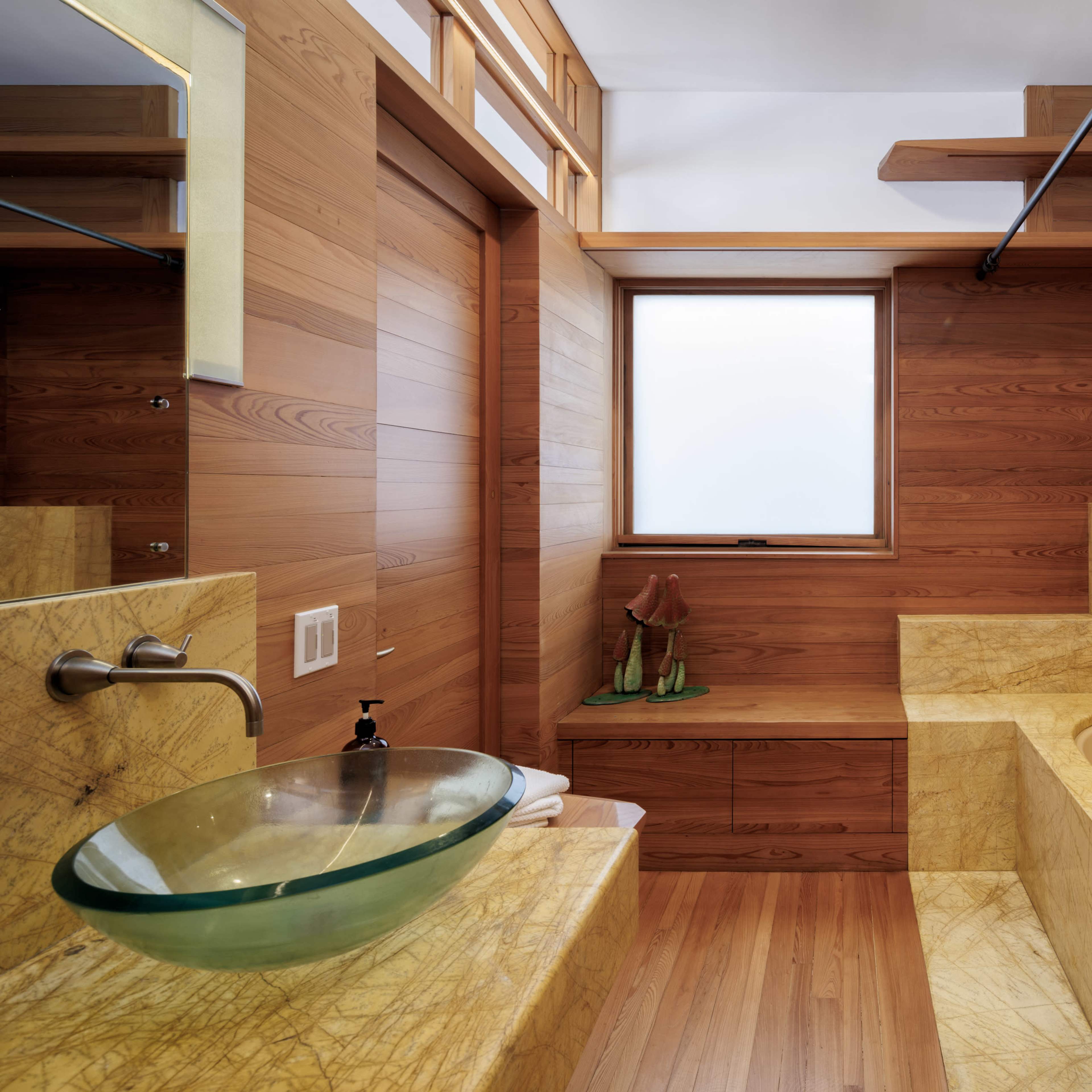 The bathroom features wooden walls, a curved tub, and a glass sink, with natural light coming through a window.