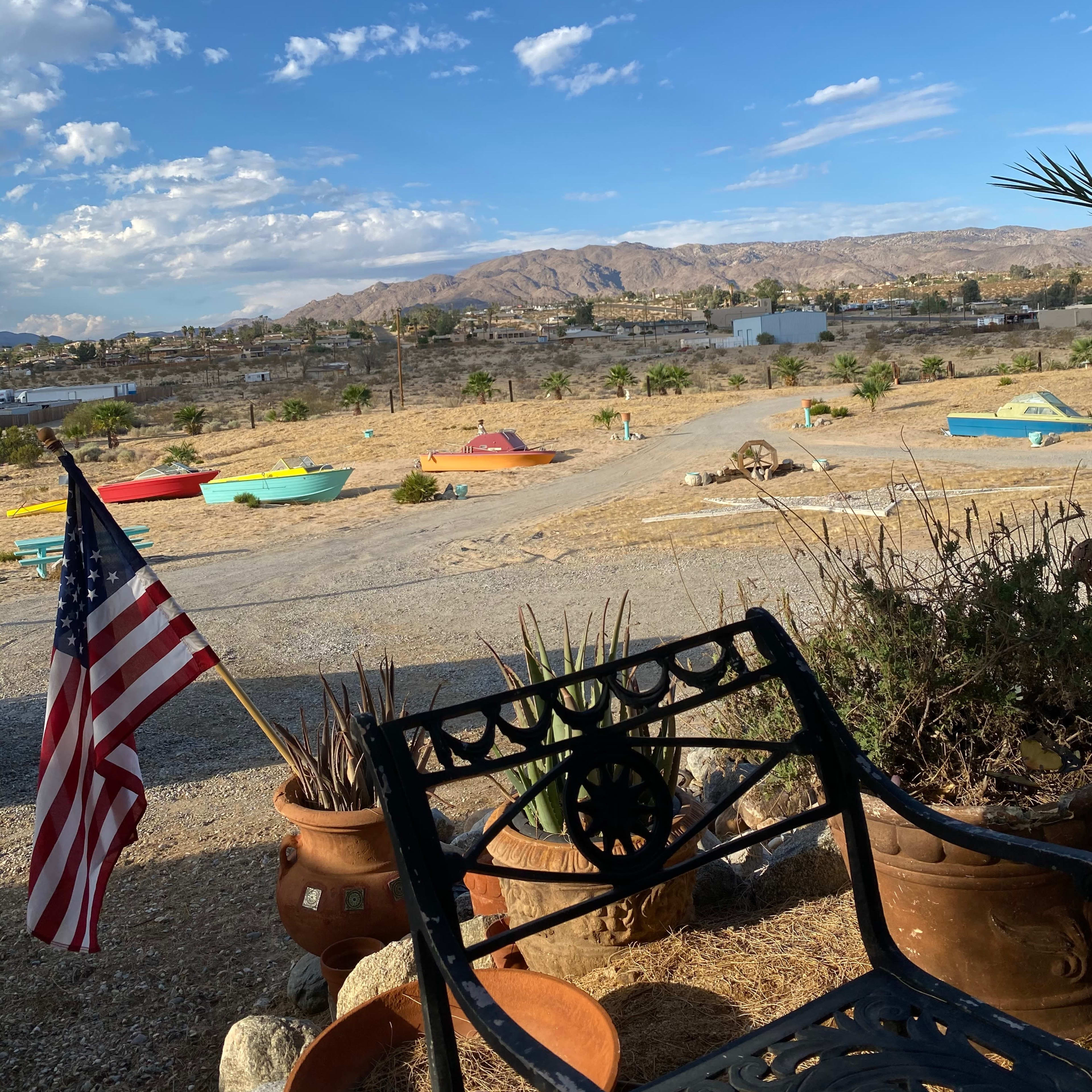 A bench with an American flag is positioned in the foreground, overlooking a landscape with boats and mountains under a partly cloudy sky.