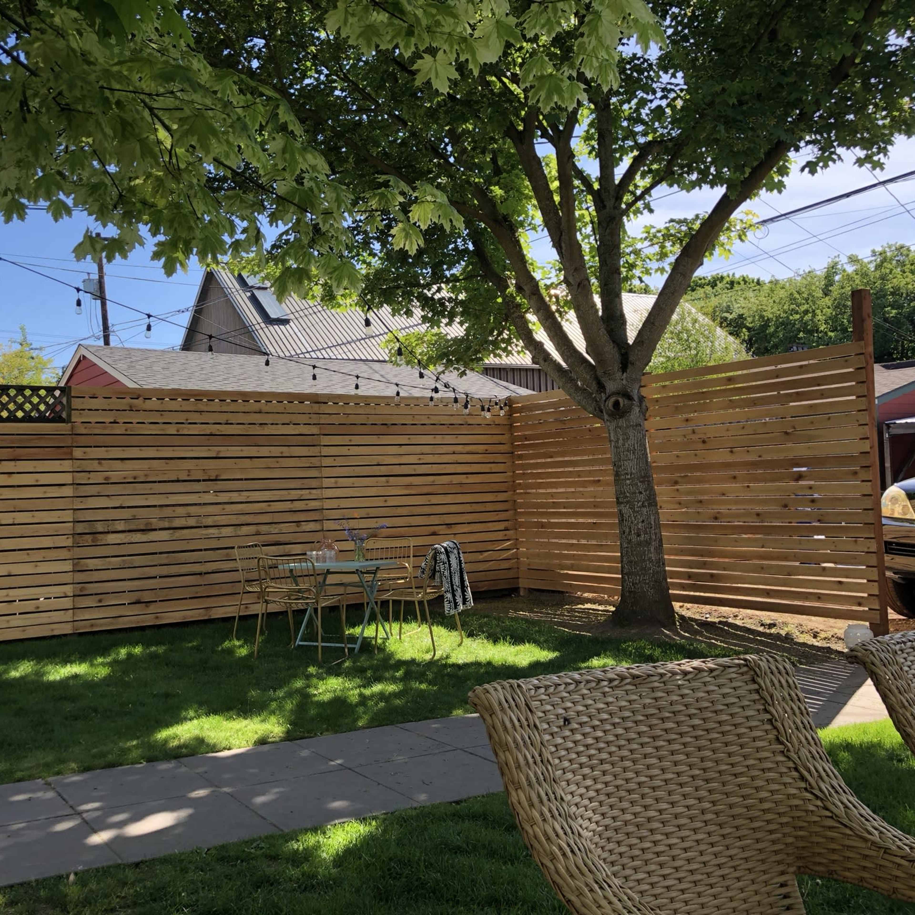 A fenced backyard with a grassy area, a round table and chairs, and a large tree providing shade.