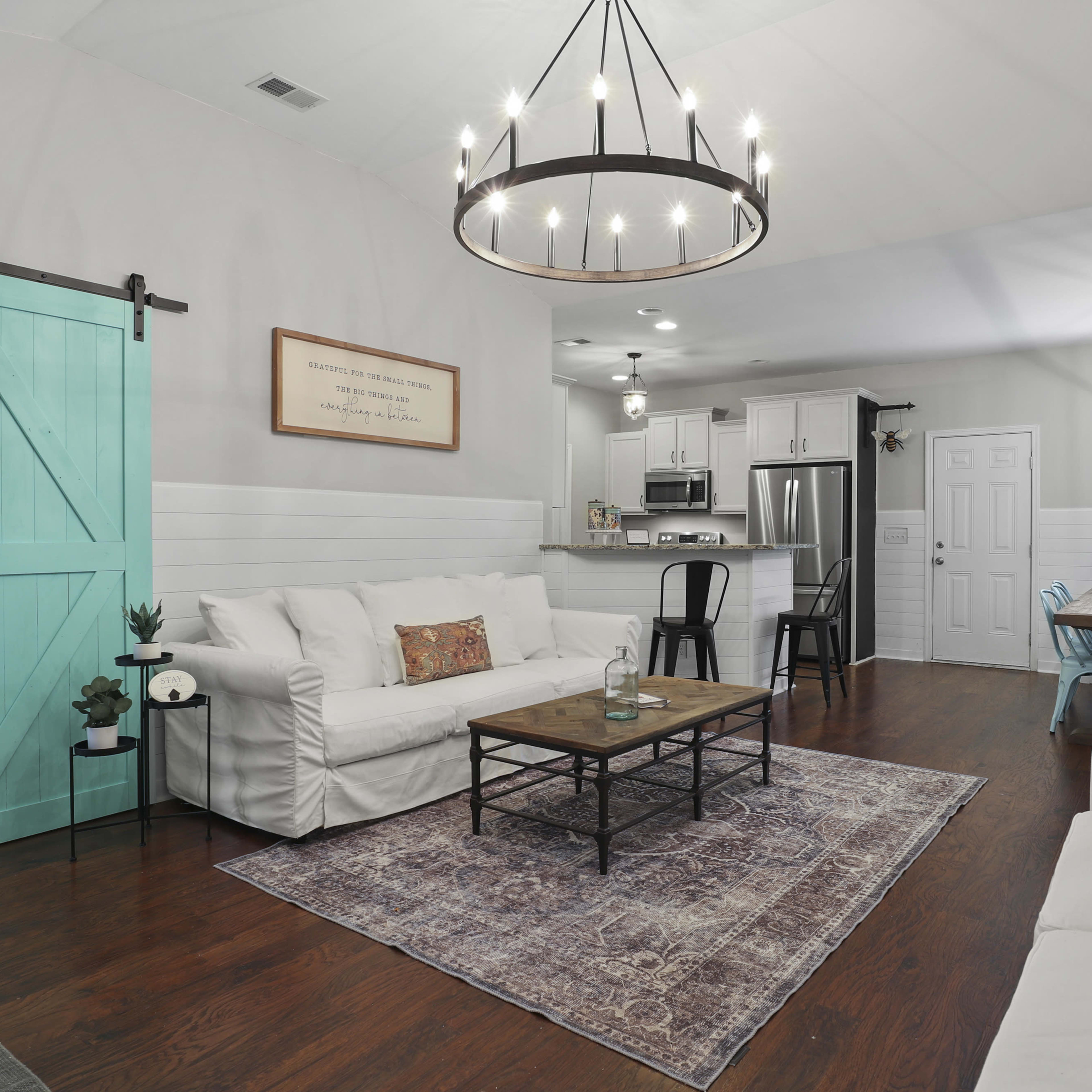 A cozy living room features white sofas, a wooden coffee table, and a kitchen with modern appliances, all set against a backdrop of light-colored walls and dark wooden flooring.
