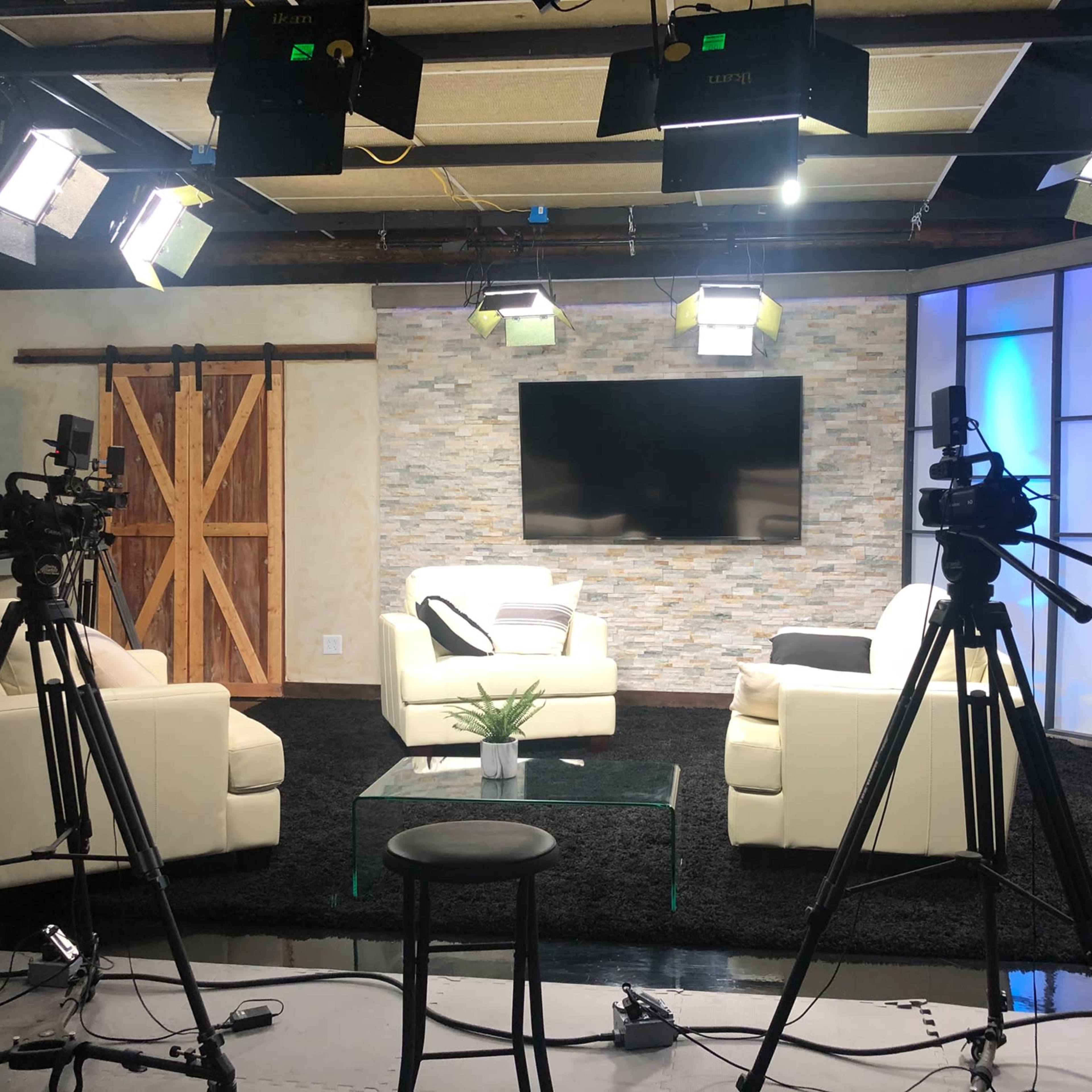 A television studio setup features two white sofas, a glass coffee table, and multiple cameras positioned around a well-lit set with a stone accent wall and a large screen.