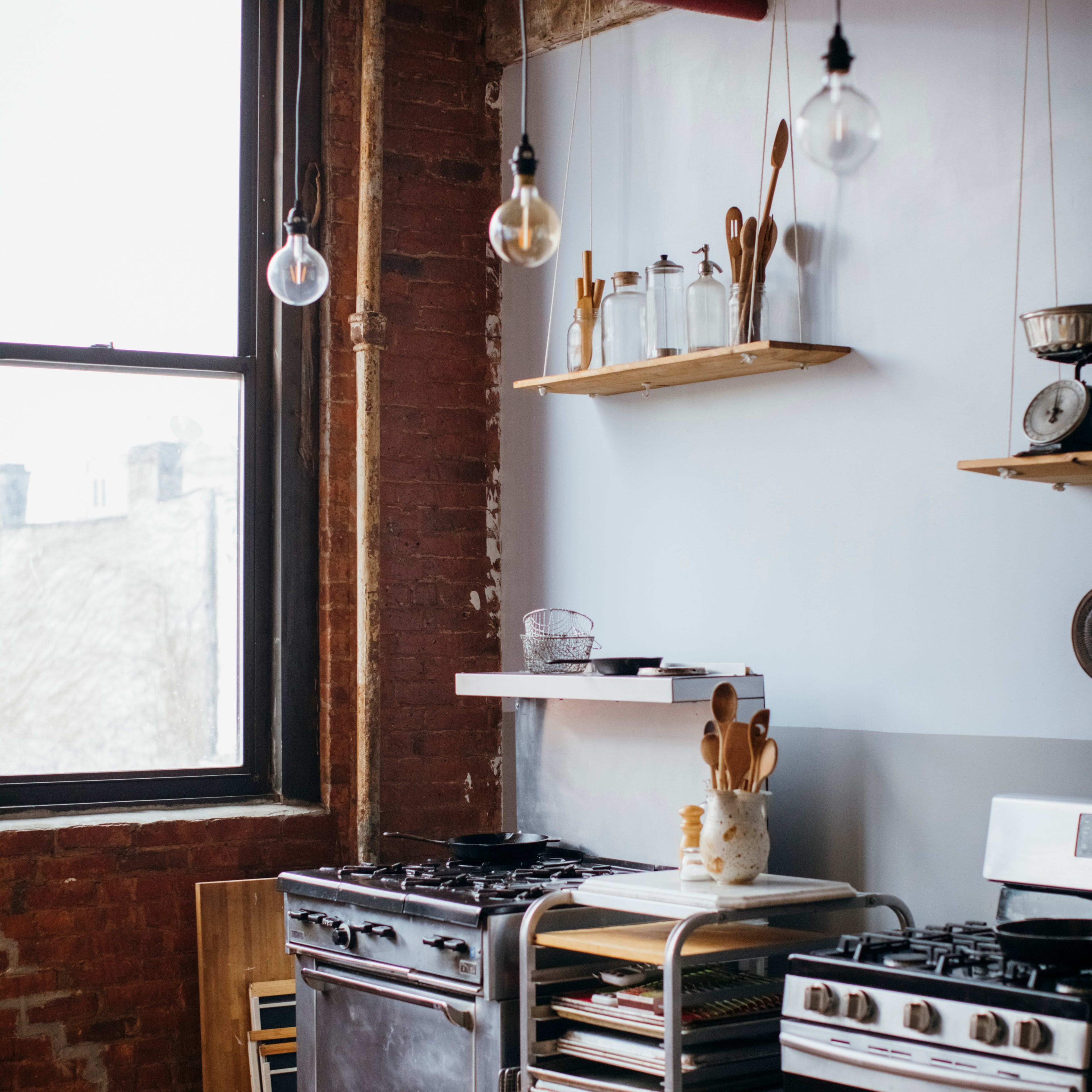 A rustic kitchen with exposed brick walls, a gas stove, a metal shelving unit, and hanging light fixtures.