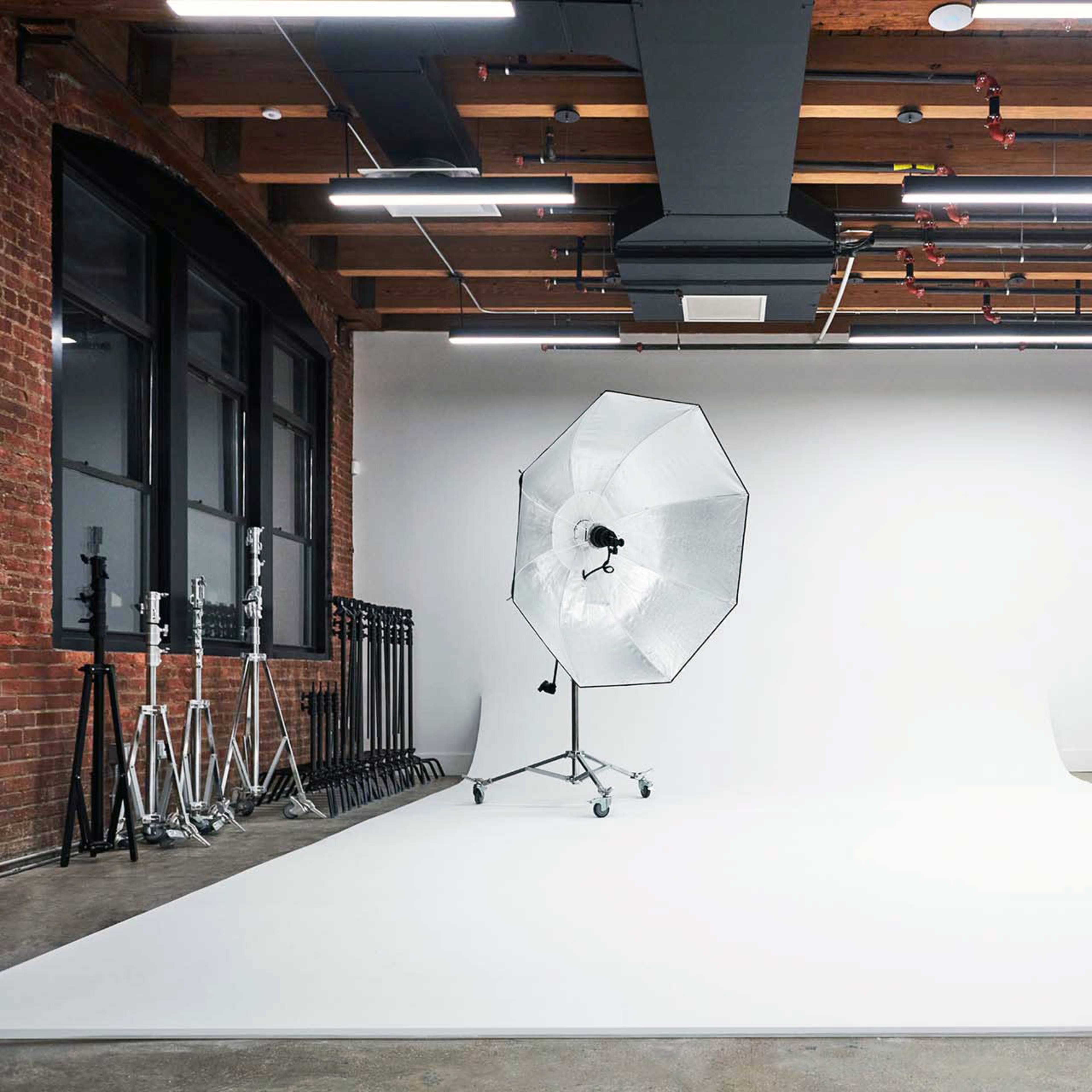 A spacious photography studio featuring a white backdrop, an umbrella softbox on a stand, and equipment such as light stands lined against a brick wall.