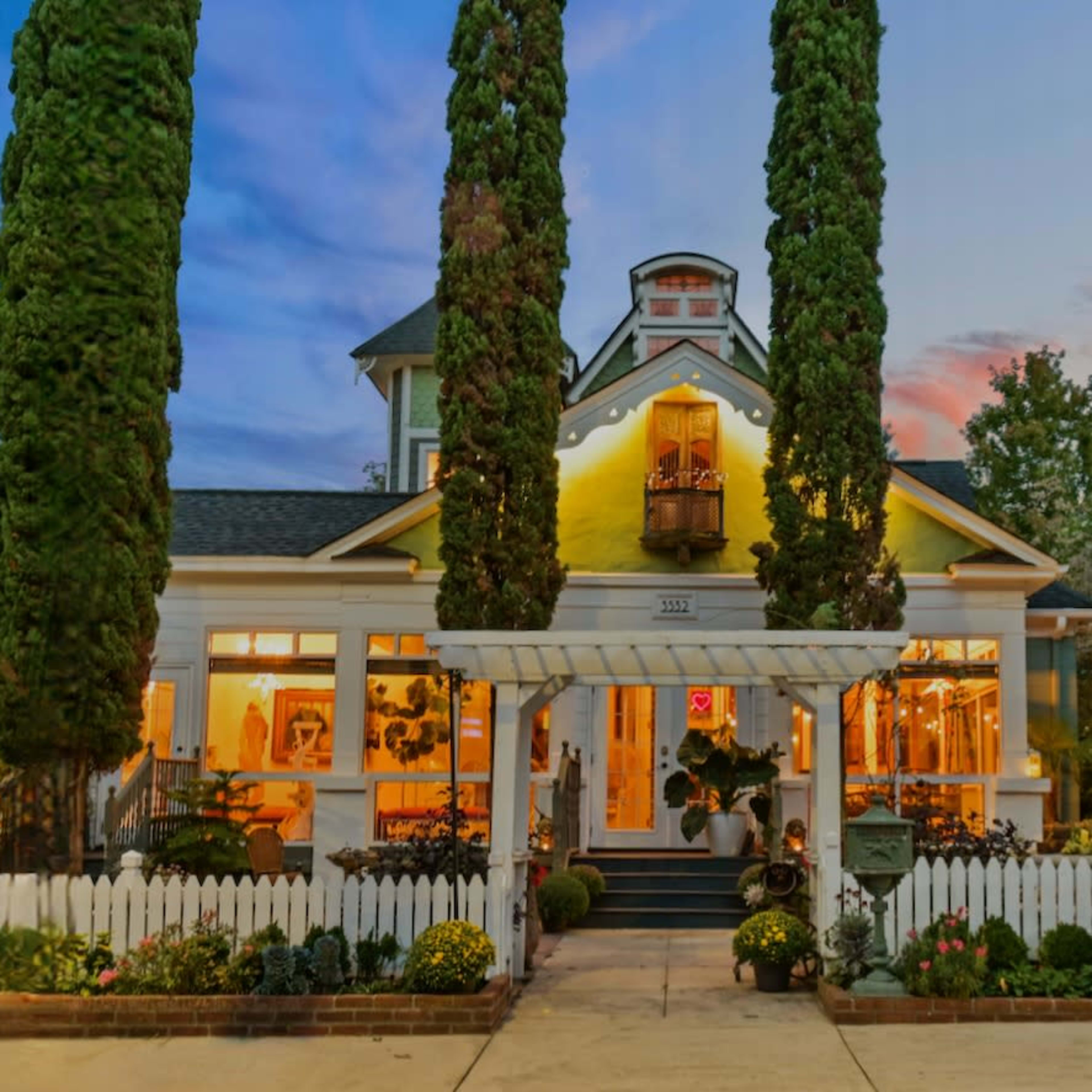 A large, white house with tall green trees in front and a picket fence is illuminated at dusk.