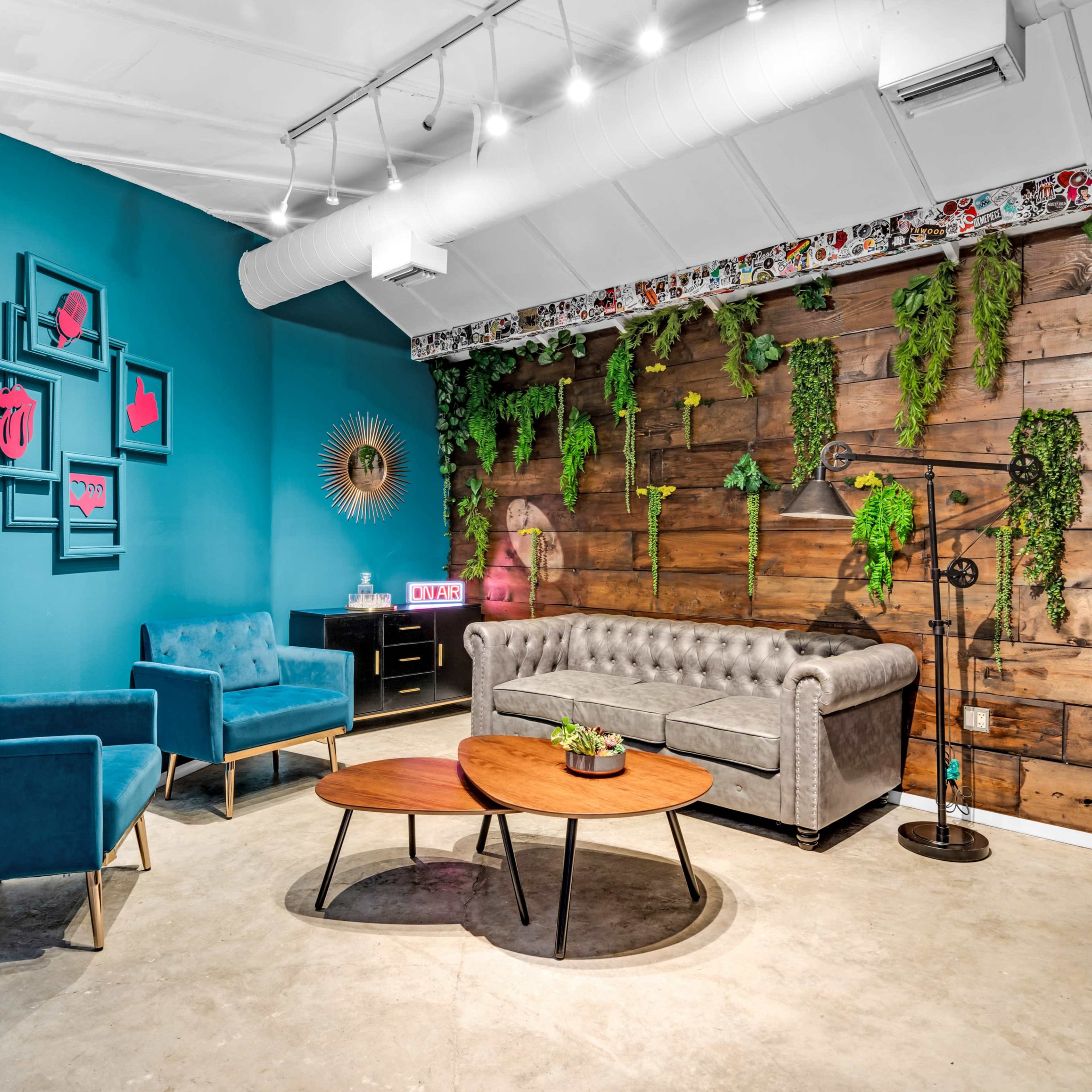 A cozy lounge area featuring a gray velvet sofa, teal chairs, wooden accent walls, and plants hanging from the ceiling.
