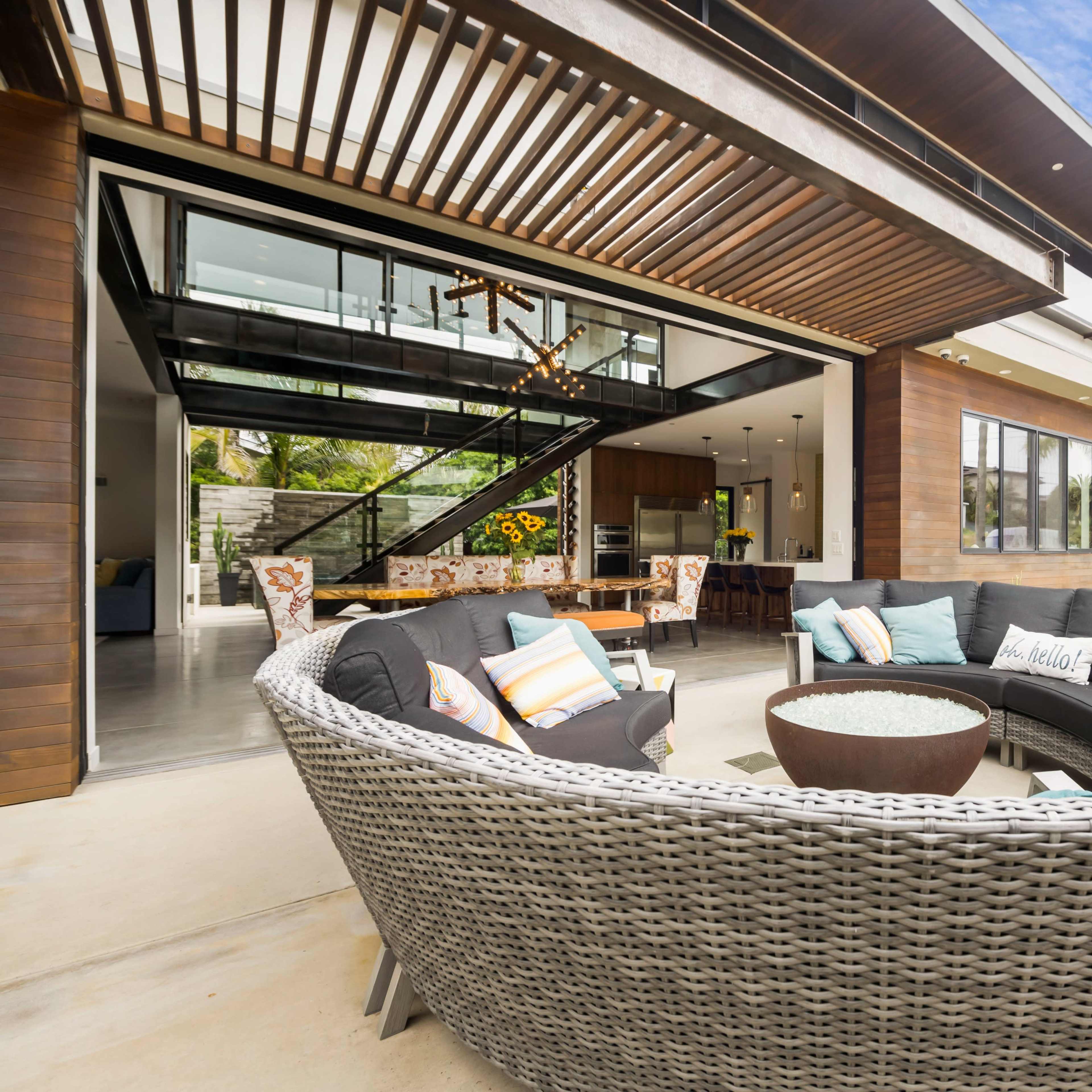 A modern outdoor seating area with a circular arrangement of wicker chairs surrounding a fire pit, adjacent to a spacious glass-fronted house.