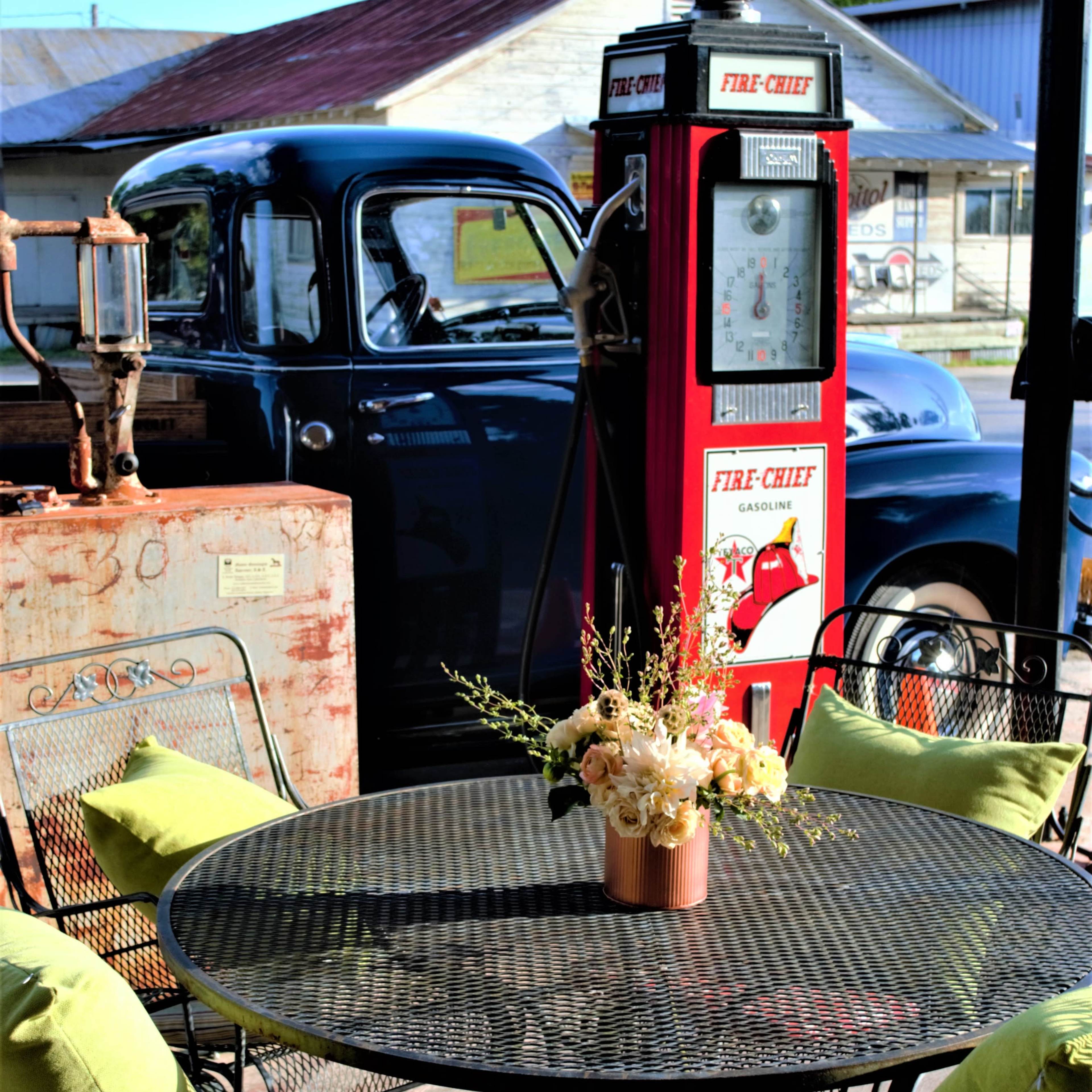 The scene features a table set with decorative cushions and a floral arrangement, alongside an antique gas pump and a vintage truck in the background.