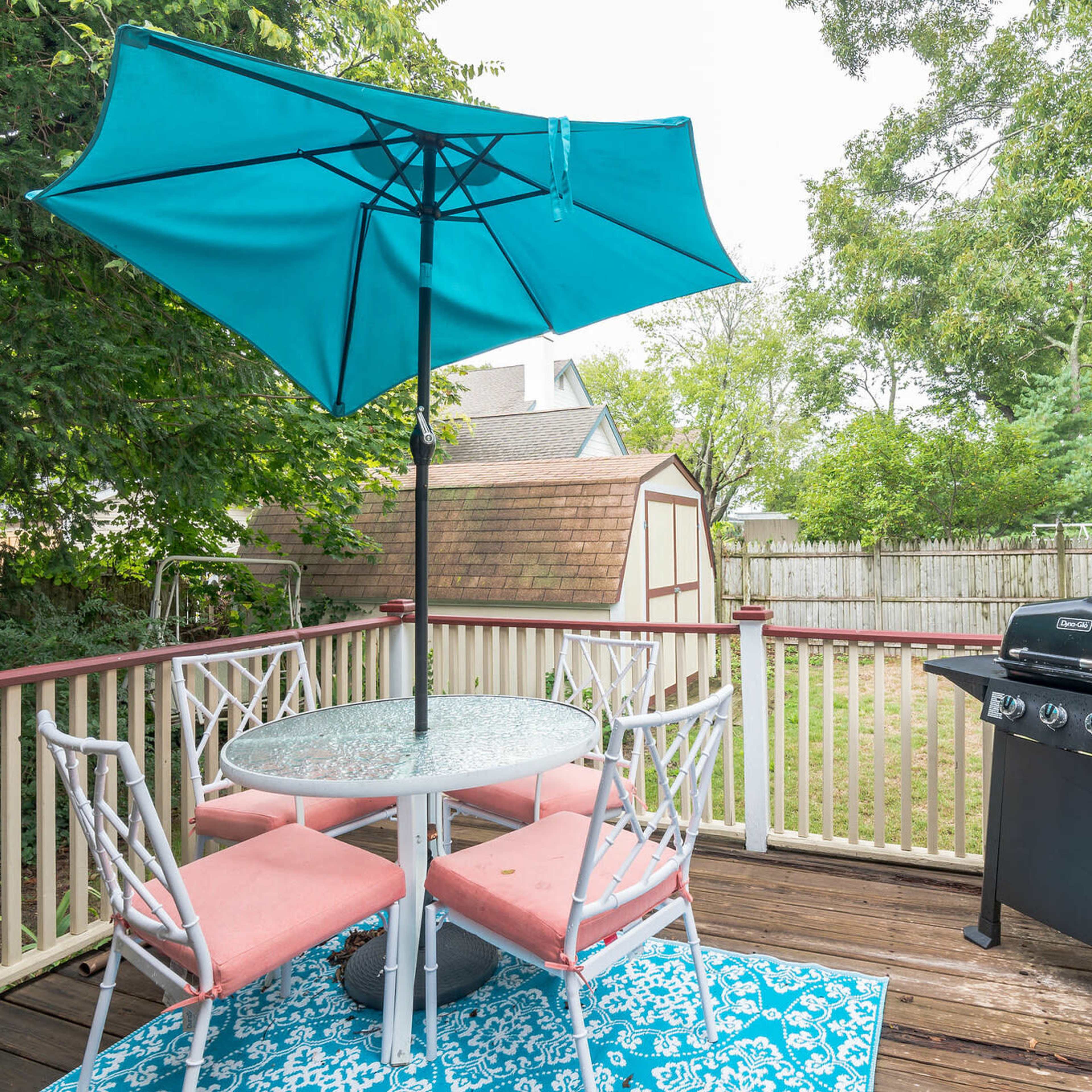 A deck features a round table with pink chairs, an umbrella, and a grill, surrounded by greenery and a shed in the background.