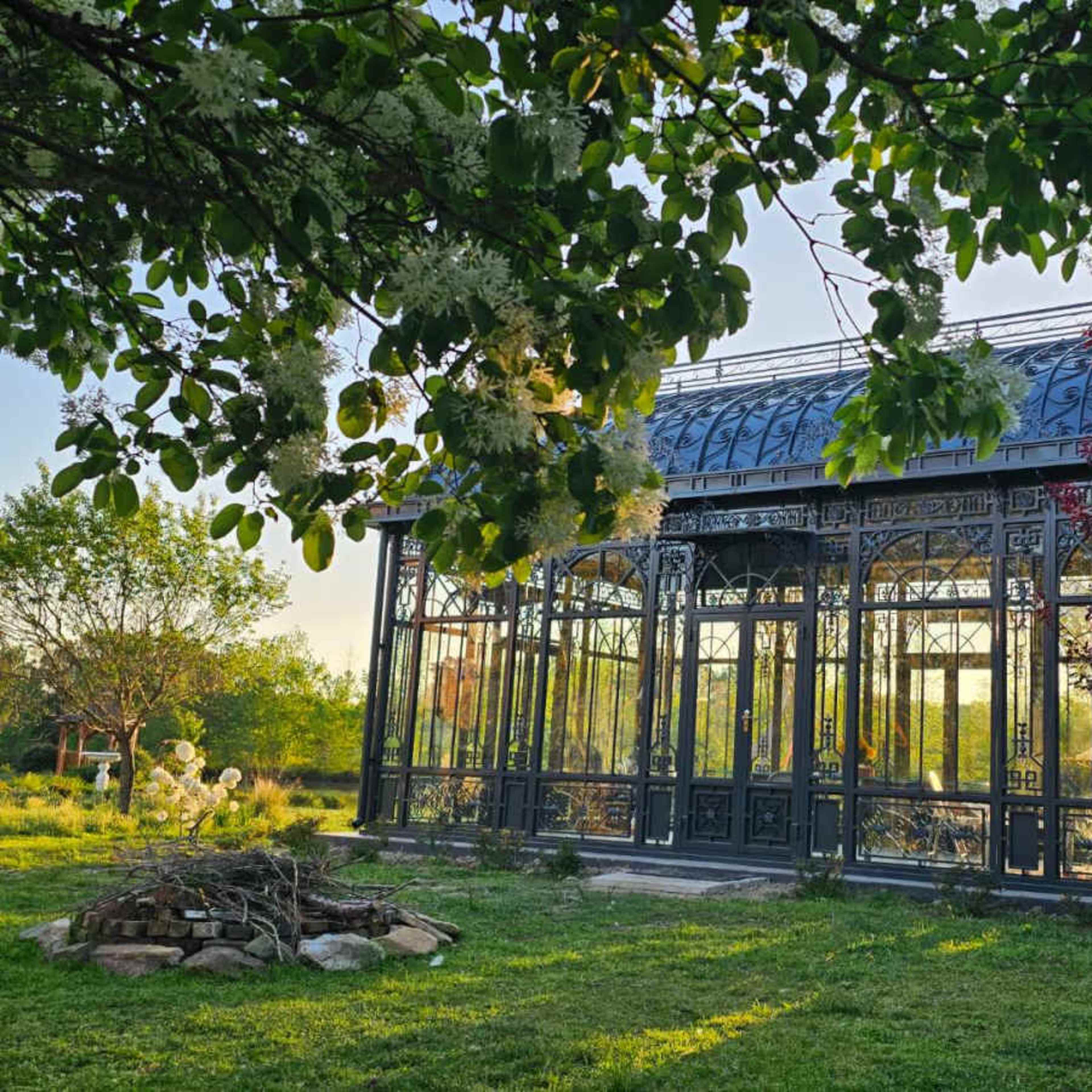 A metal-framed greenhouse surrounded by greenery and a stone arrangement in a garden.