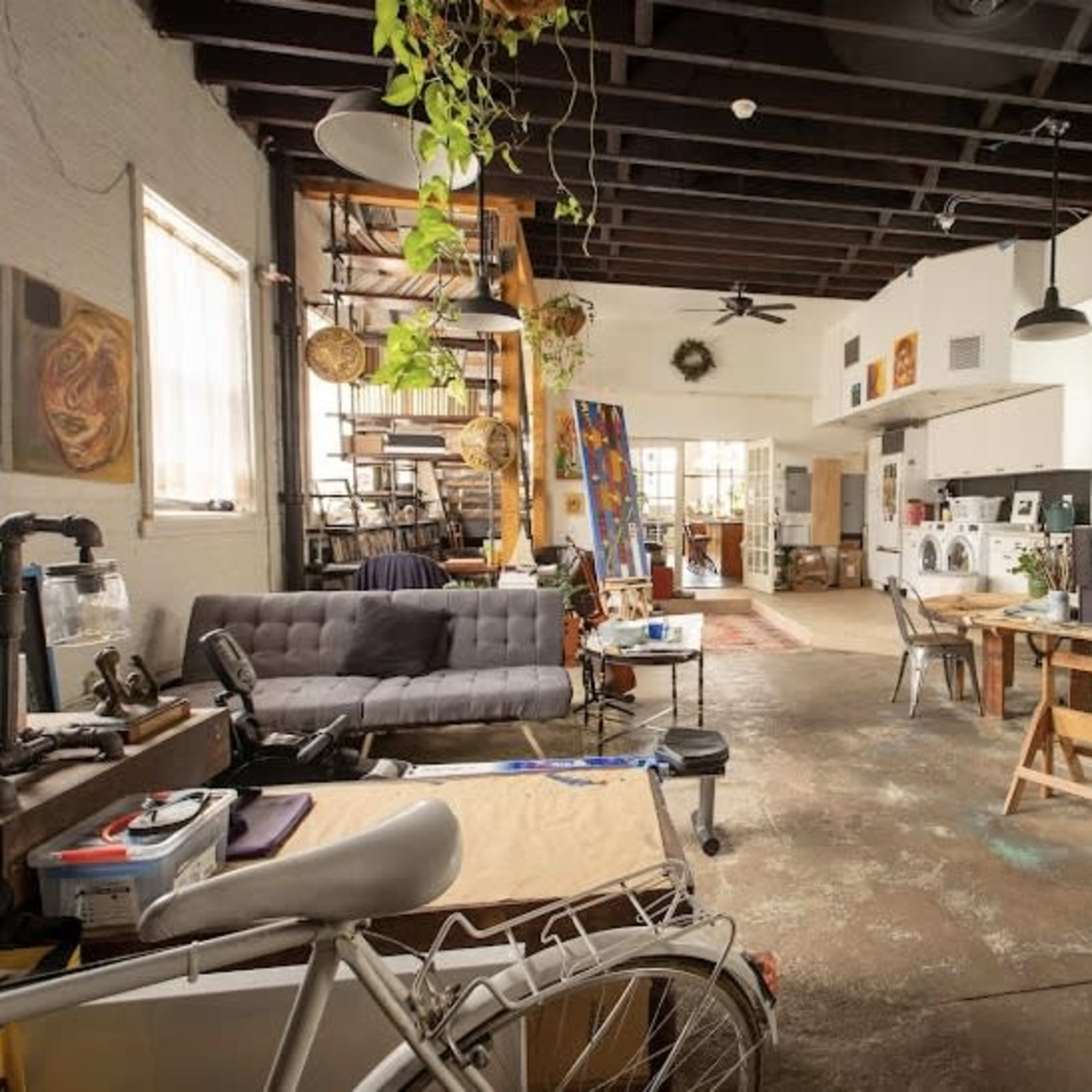 A spacious artist's studio featuring workstations, a sofa, and large windows with various artworks displayed on the walls.