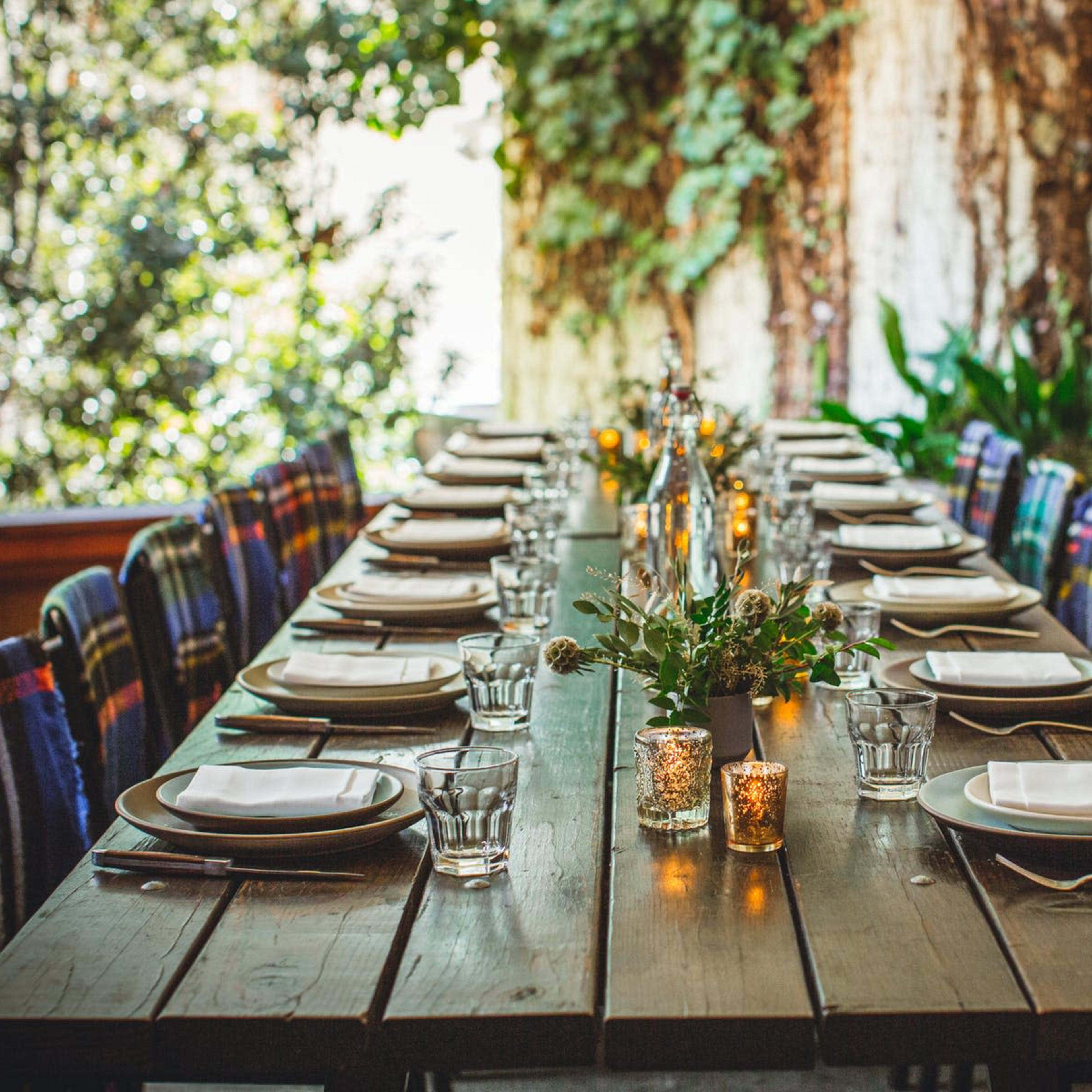 A long wooden table is set for a meal, featuring plates, glasses, and small candles, surrounded by vibrant greenery.