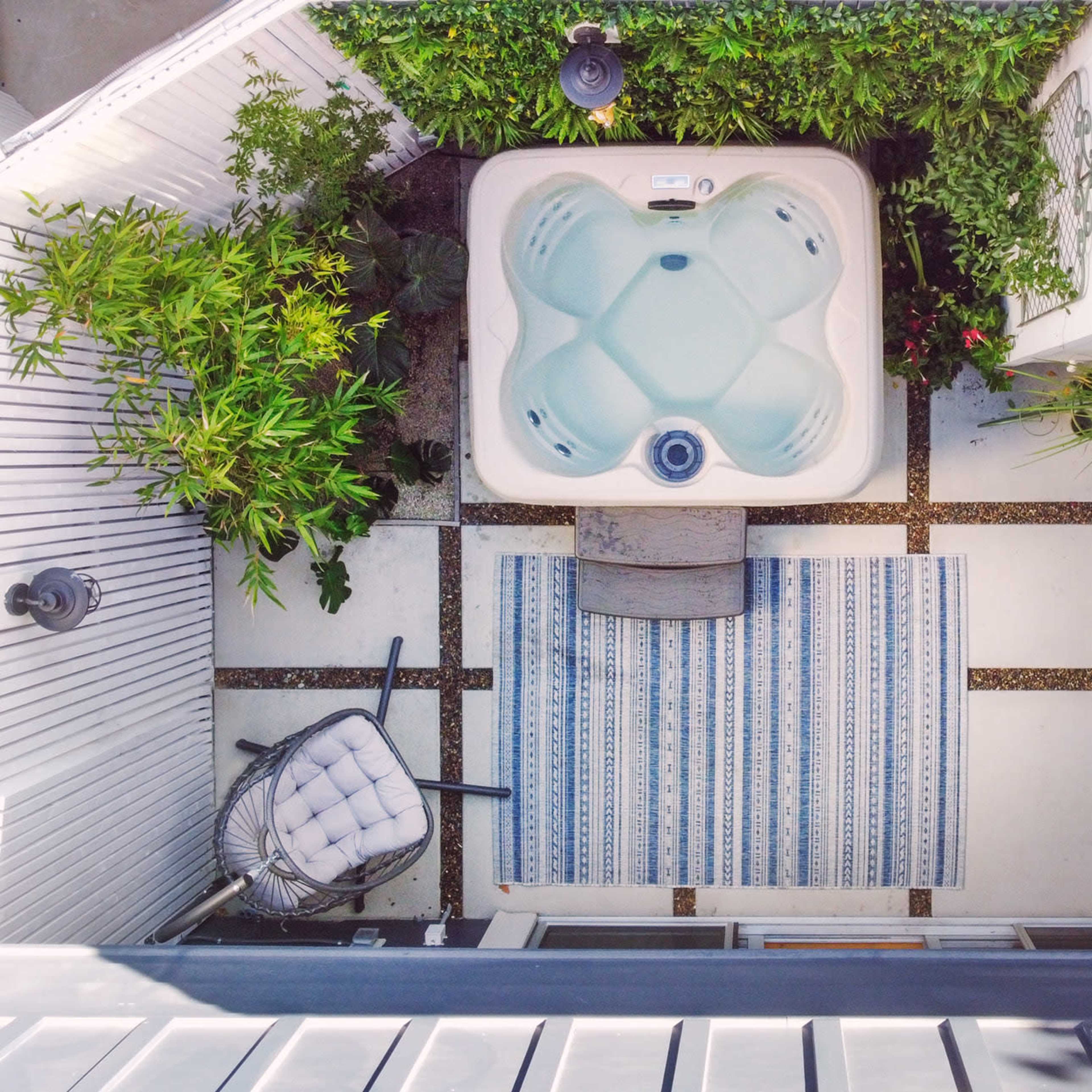 An outdoor patio with a hot tub, a patterned rug, and seating arrangements surrounded by plants.