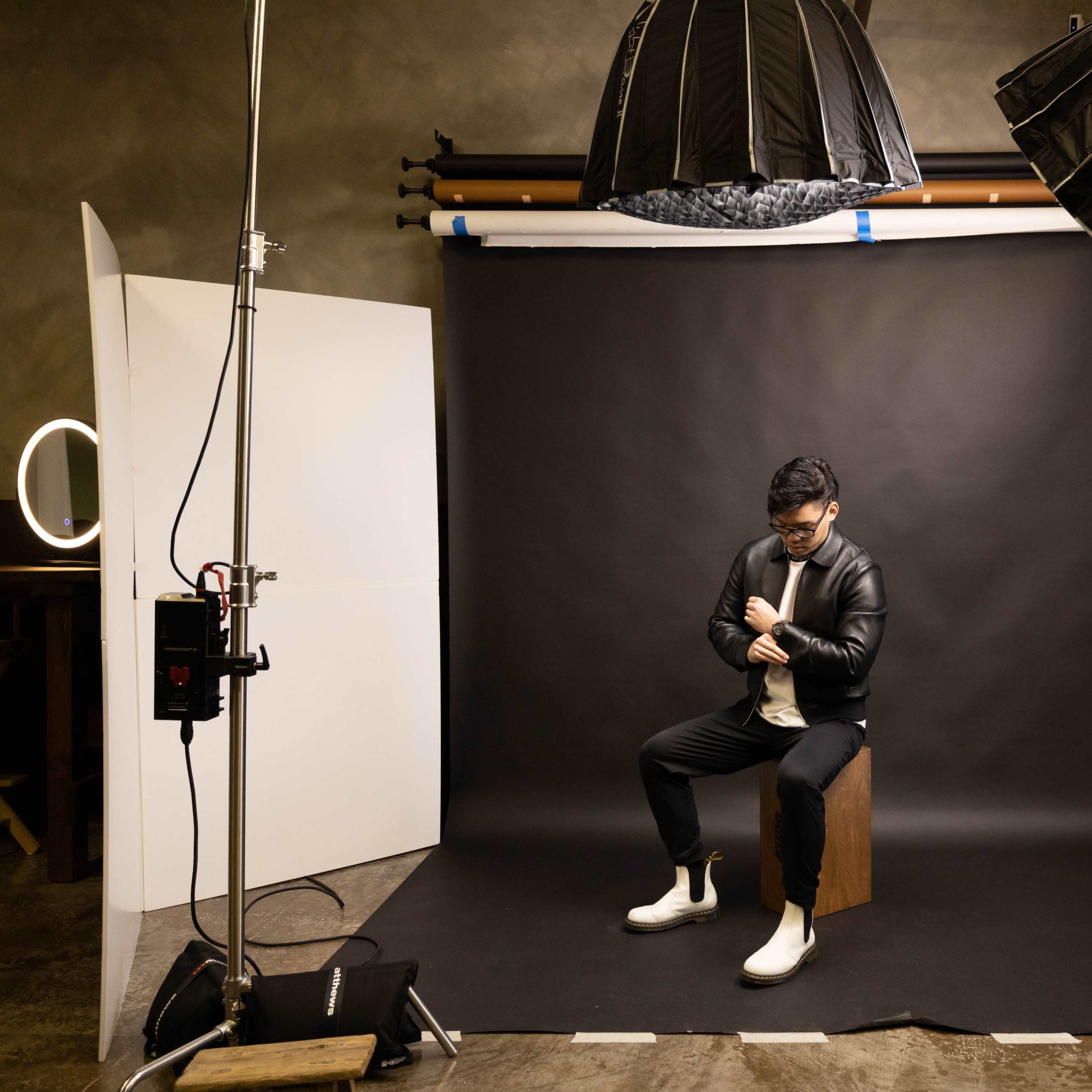 A man wearing a black jacket and white shoes sits on a wooden box in a photography studio, surrounded by lighting equipment and a black backdrop.