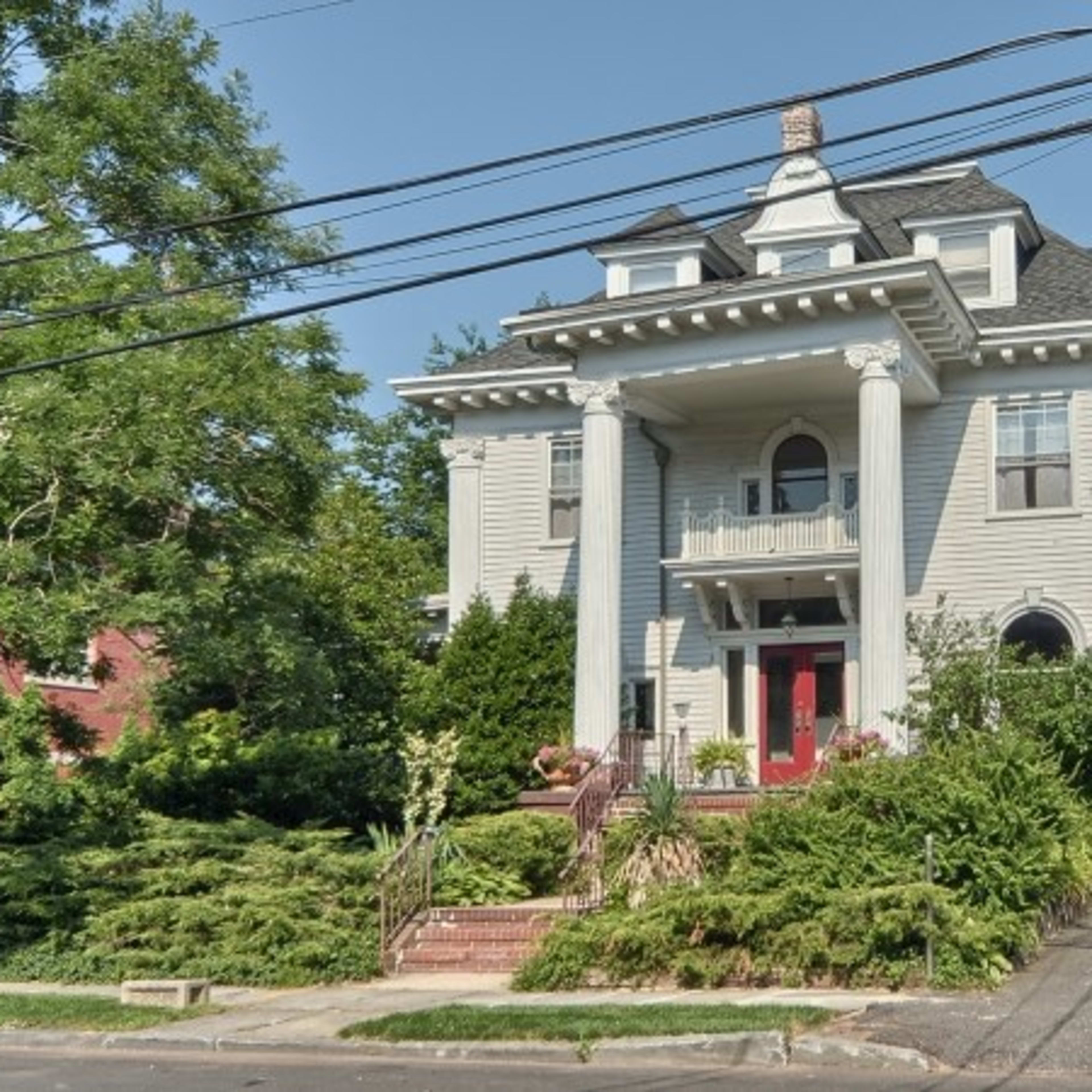 A large, multi-story house with white siding and a prominent porch is surrounded by trees and greenery, with power lines overhead.